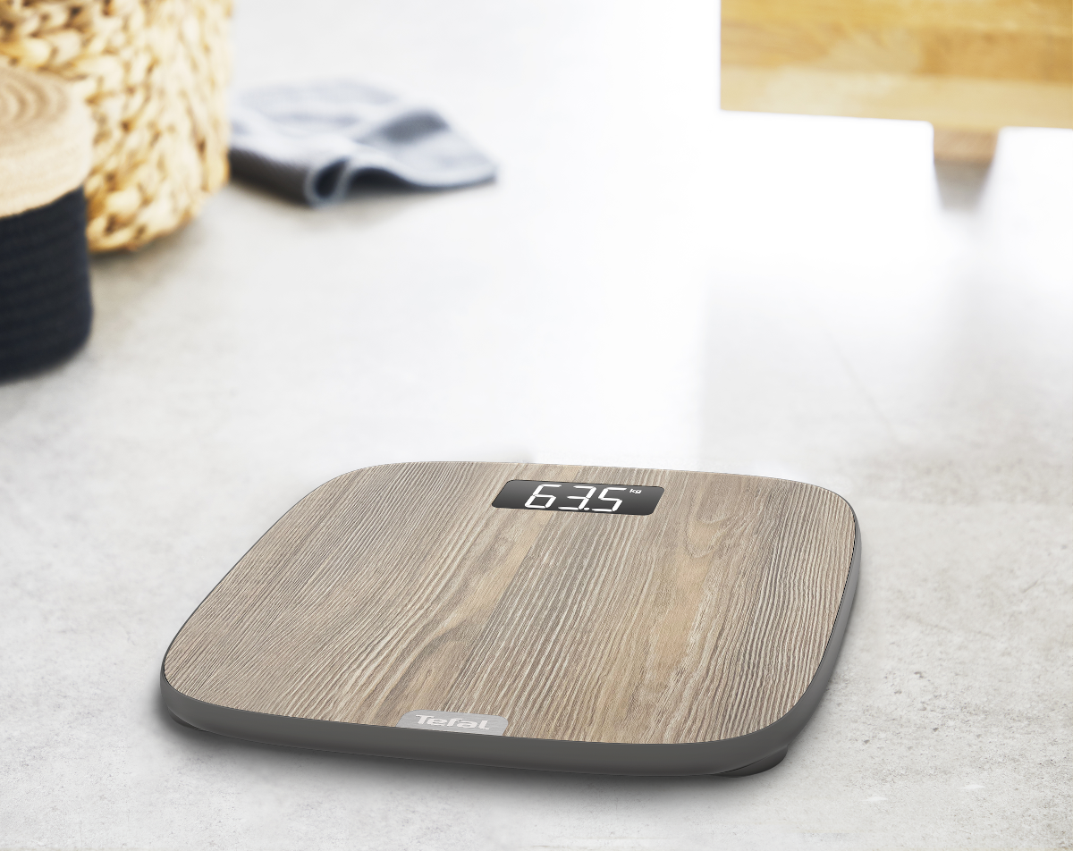 Een digitale weegschaal met houten textuur toont 63,5 kg. Een mand en een handdoek bevinden zich op de wazige achtergrond.