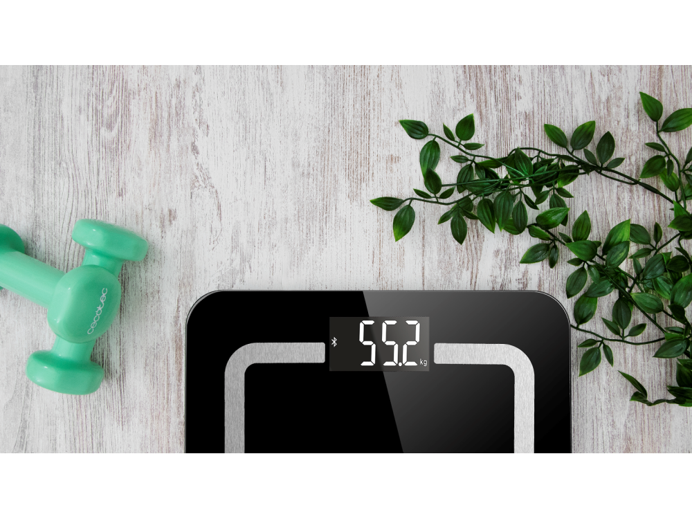 Báscula inteligente negra que muestra 55,2 kg, con una pesa verde y planta.