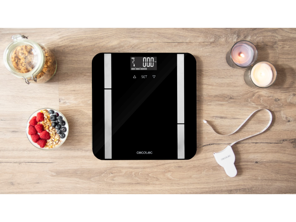 Báscula digital negra con detalles blancos sobre una mesa de madera. Yogur con bayas, granola y una cinta métrica cerca.