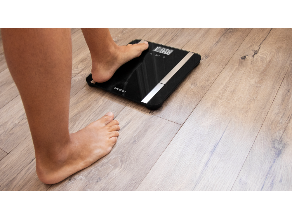 Una persona de pie en una báscula digital negra sobre un suelo de madera. Un pie está en la báscula, el otro al lado.
