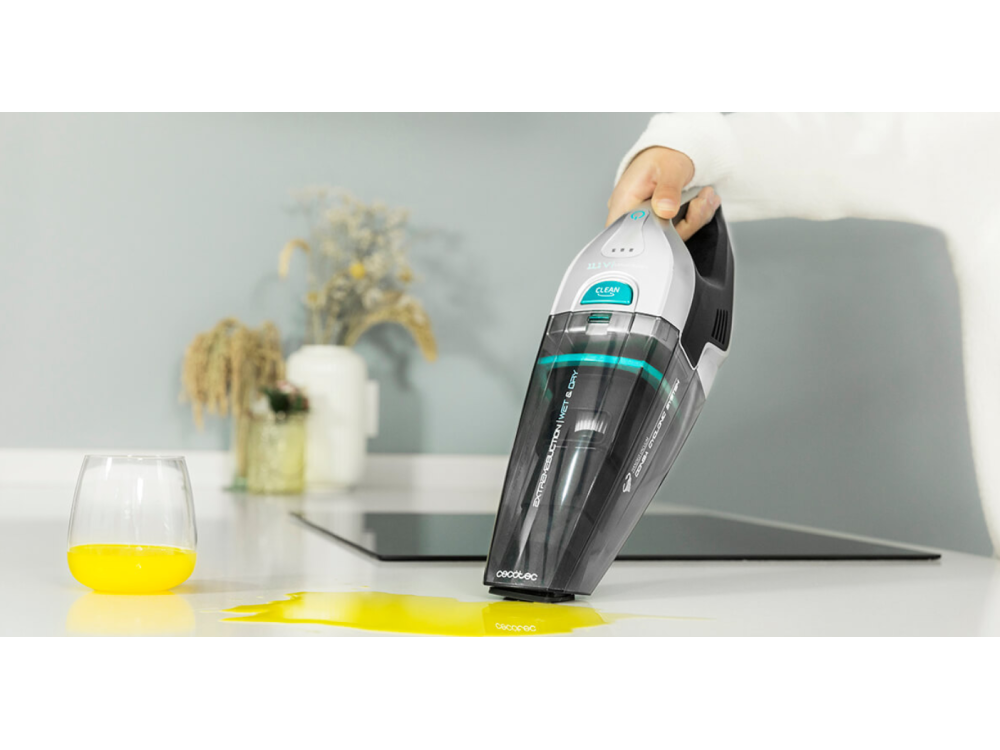 Una aspiradora de mano limpia un derrame amarillo. Hay un vaso y otros objetos en el fondo.