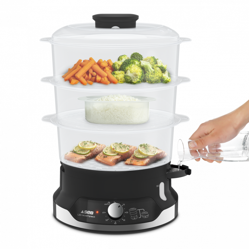 Steamer with food in transparent containers. Hand pouring water. Vegetables, fish, and rice are inside.