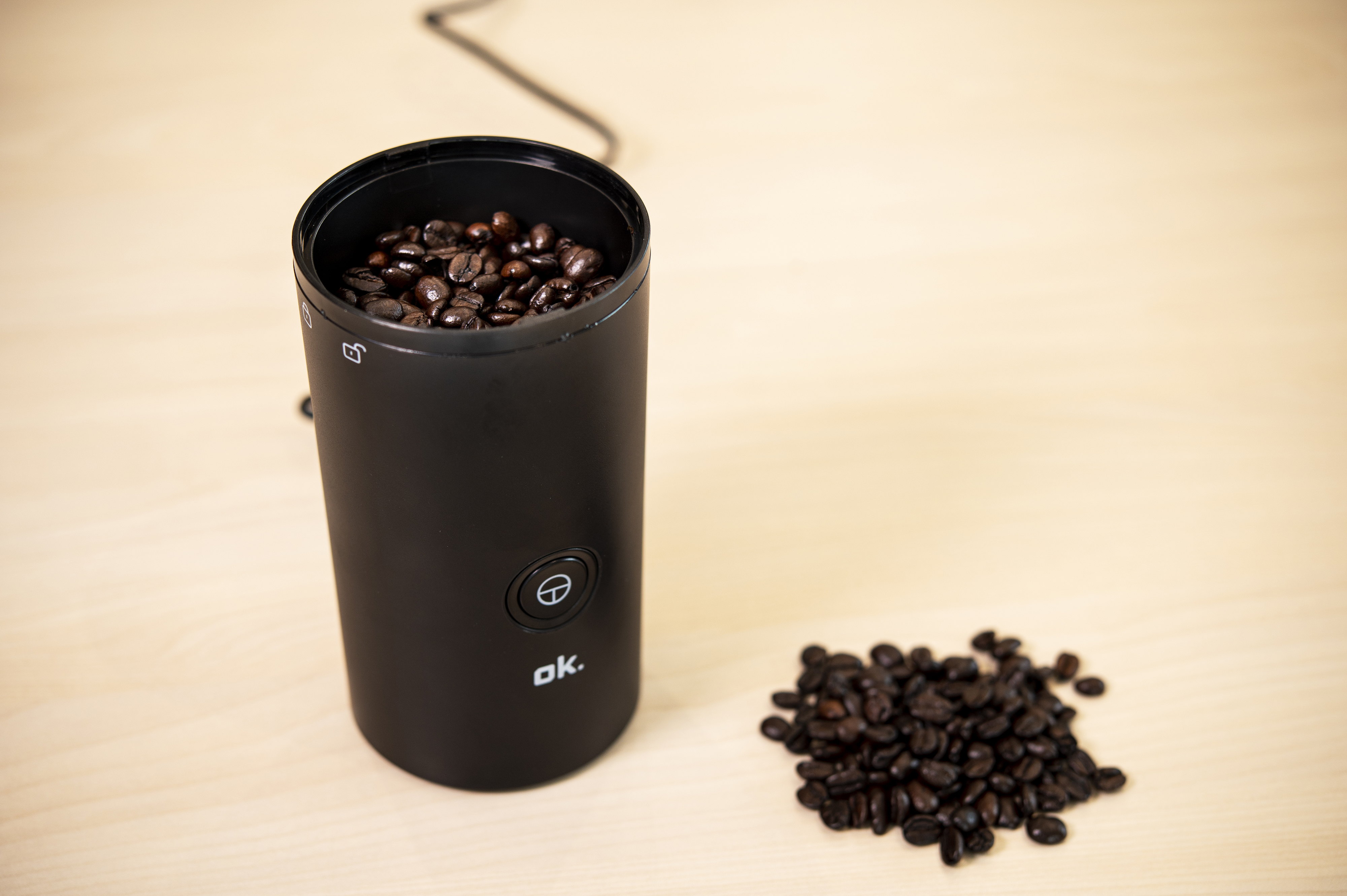 A black coffee grinder filled with coffee beans, and a pile of beans next to it on a wood surface.