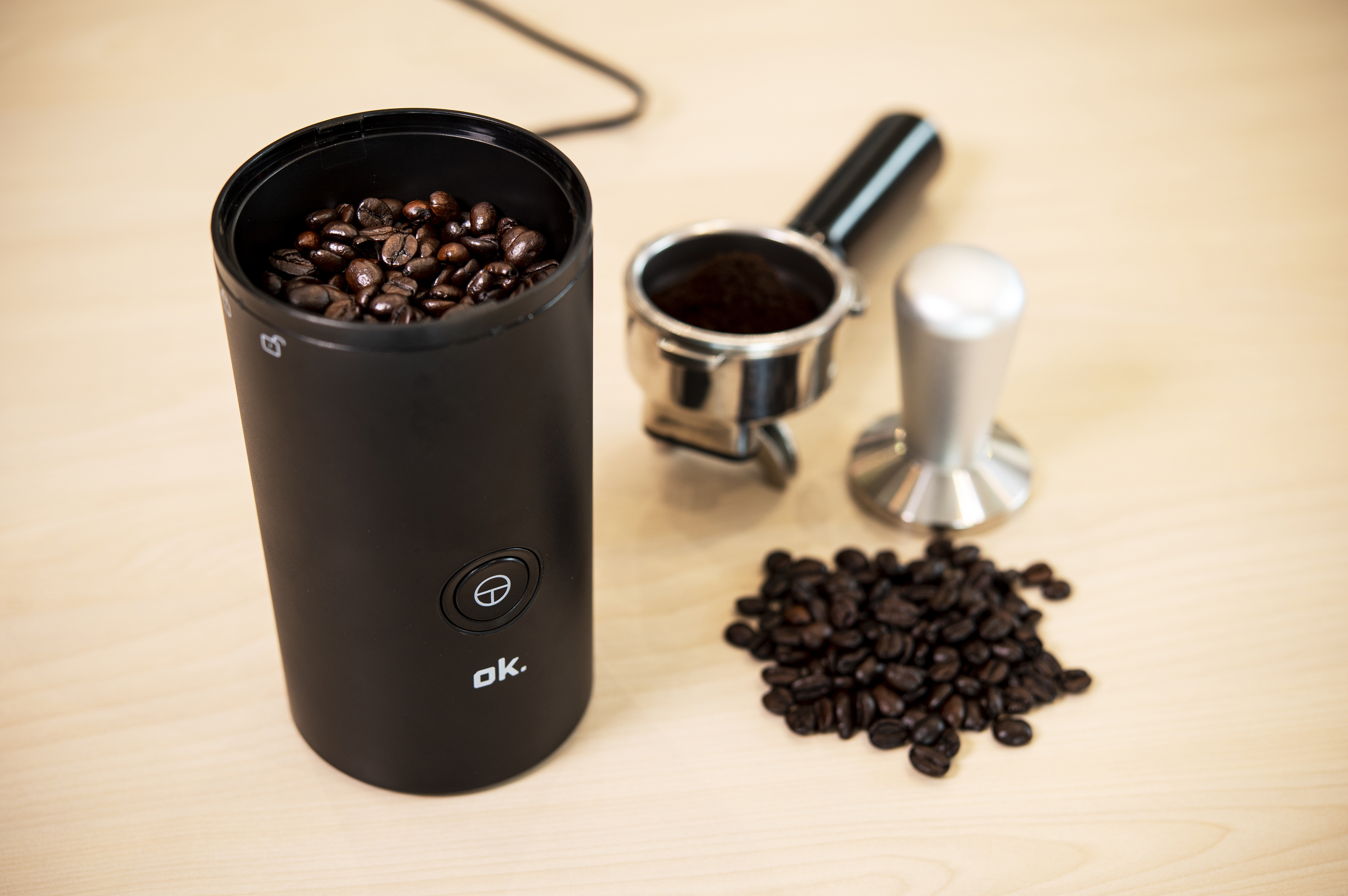 Coffee beans in a grinder, with ground coffee, portafilter, and tamper on a table.