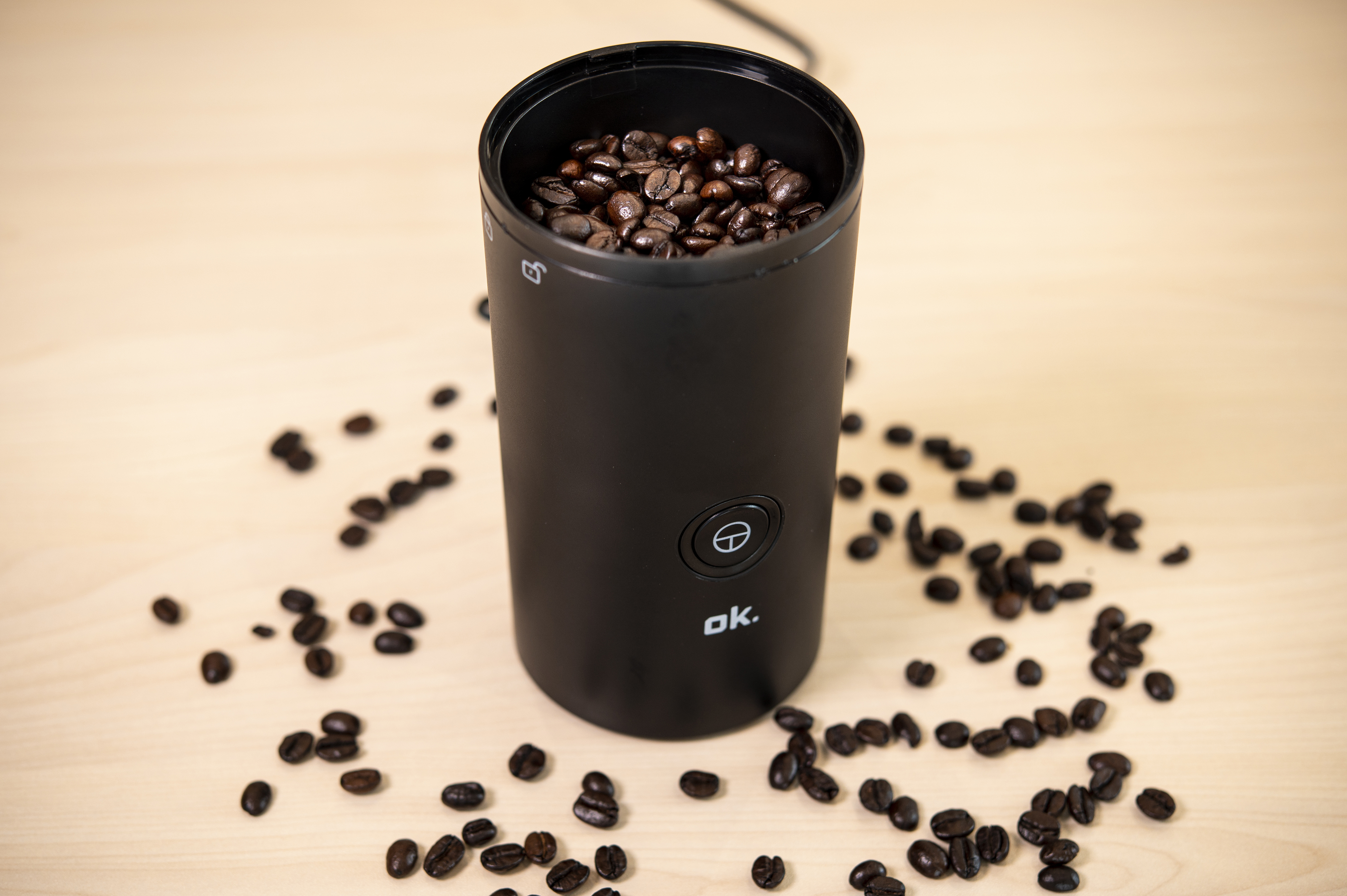 Black coffee grinder filled with beans on a light wooden table.