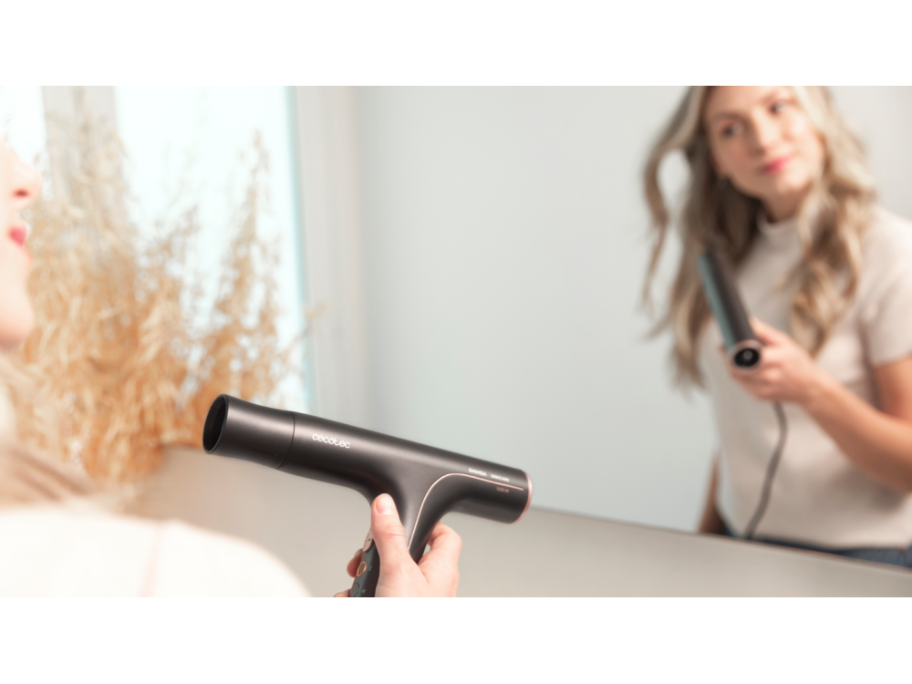 Mujer usando un secador de pelo frente a un espejo. Muestra el secador y su reflejo.