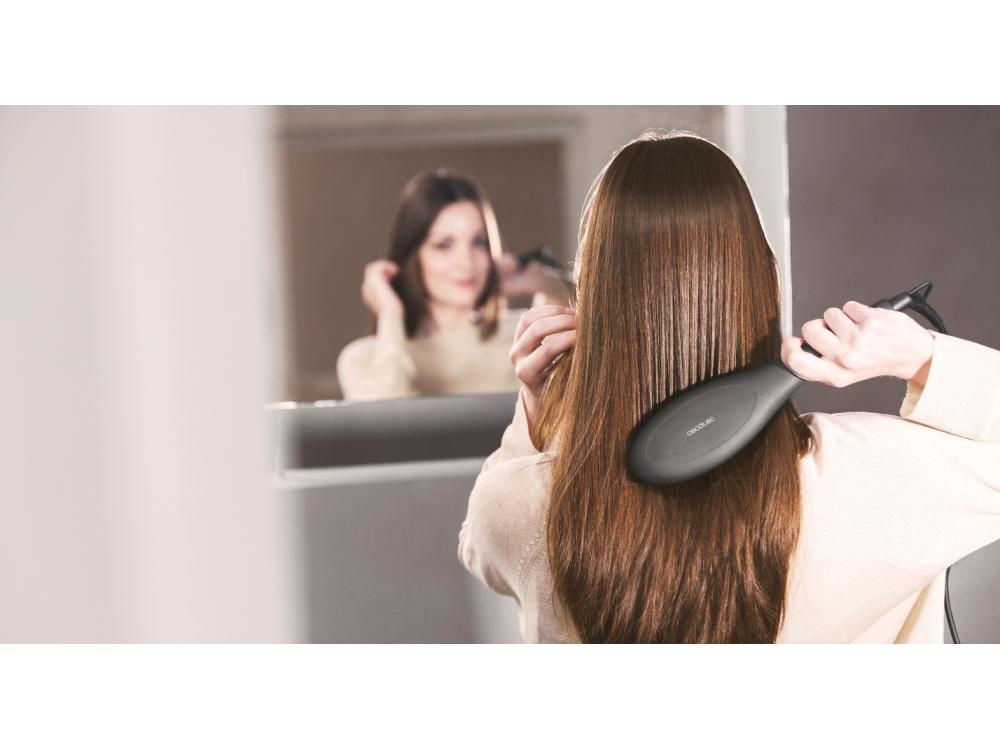 Mujer cepillando su largo cabello castaño frente al espejo, con un alisador de pelo.