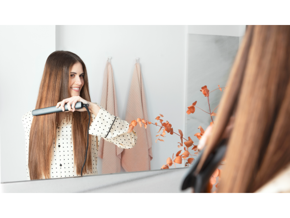 Una mujer alisándose el cabello en un espejo, sosteniendo una plancha de pelo.