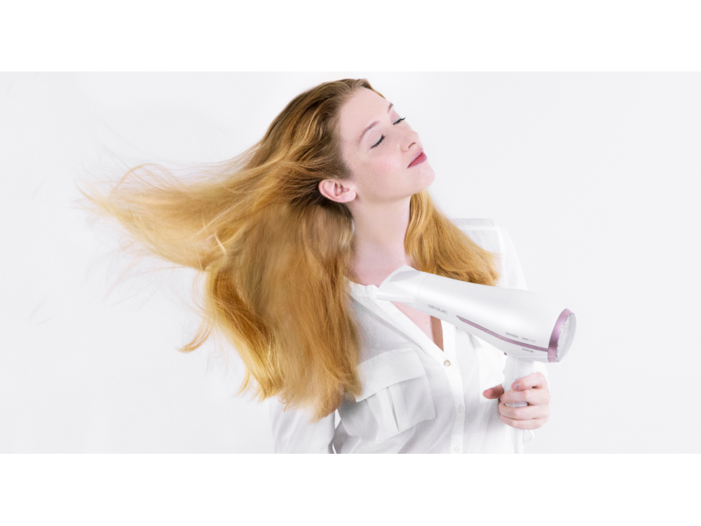 Una mujer con cabello rubio suelto se seca el pelo con un secador blanco. Lleva una camisa blanca, ojos cerrados, sobre fondo blanco.