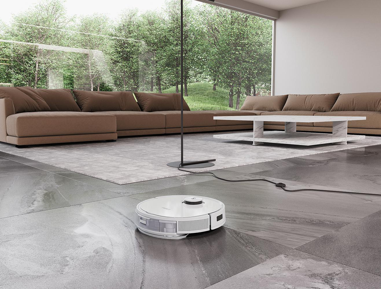Ein weißer Roboter-Staubsauger reinigt einen grauen Fliesenboden. Braune Sofas und ein weißer Tisch im Hintergrund.