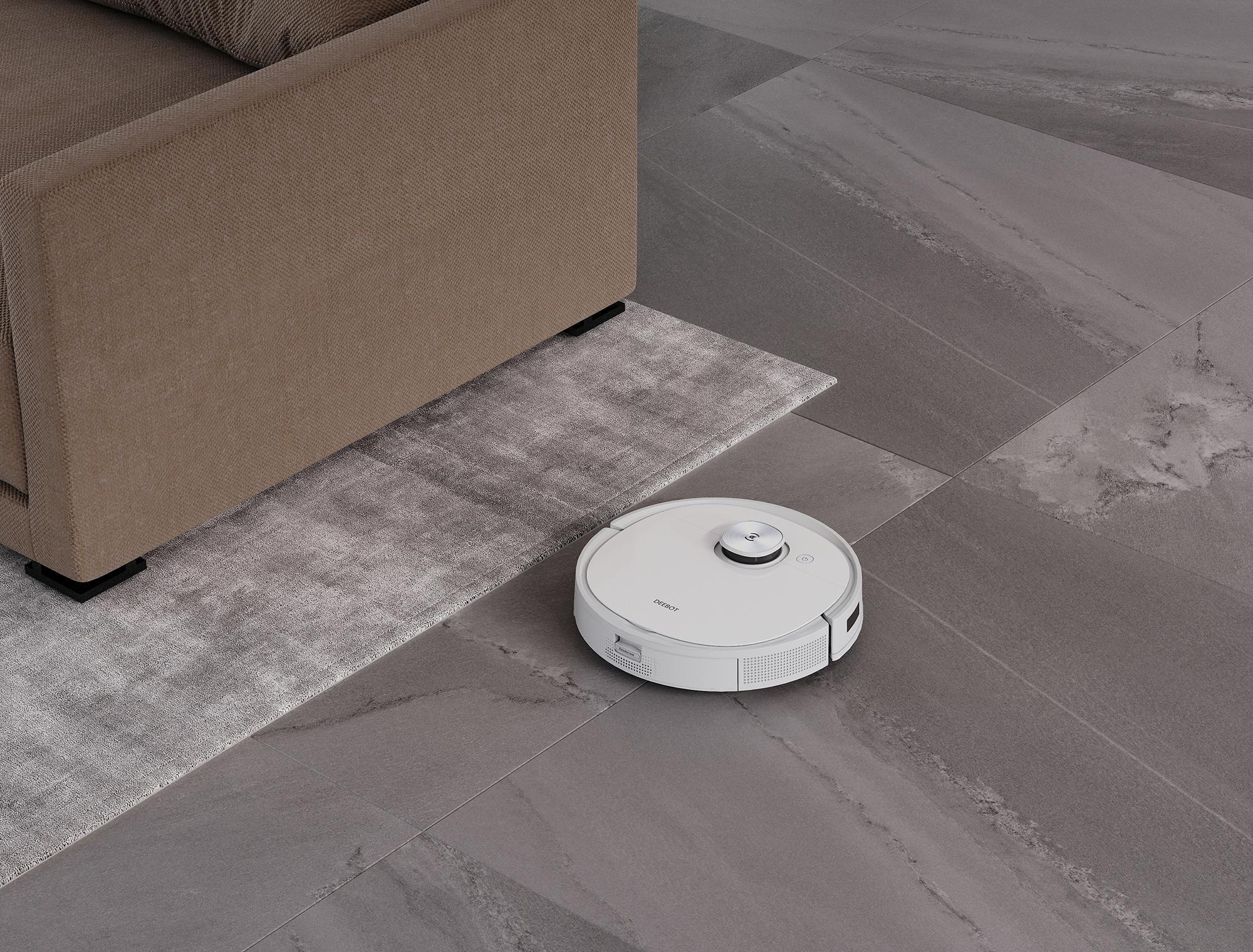 Ein weißer Roboter-Staubsauger auf einem grauen Steinboden neben einem Sofa und Teppich.