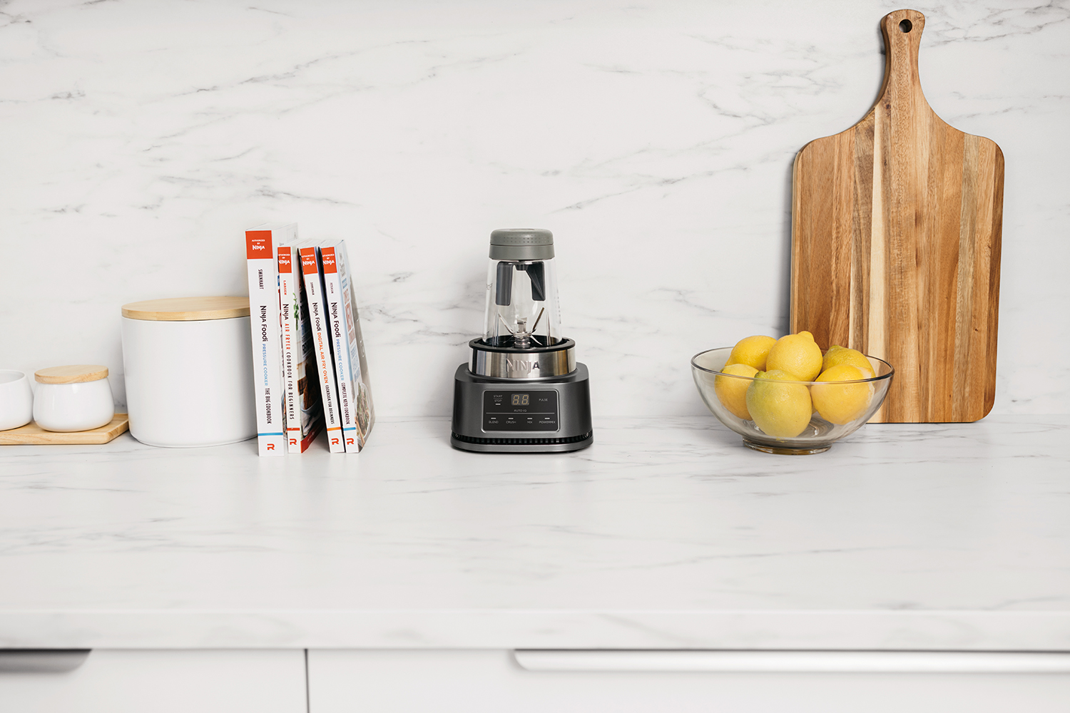 Kitchen countertop with a blender, books, lemons, and a wooden cutting board.