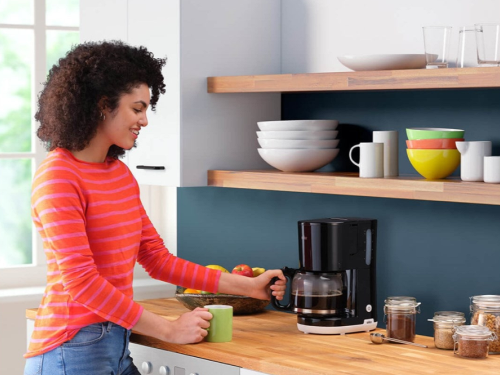 Mujer con camisa a rayas, preparando café. Ambiente de cocina con cafetera, frascos y cuencos en estanterías.