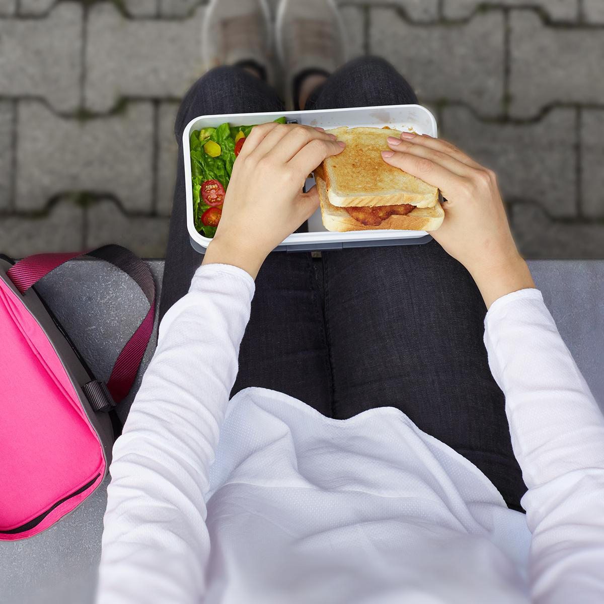 Person hält Sandwich und Salat in einer weißen Brotdose, sitzt auf einer Bank.