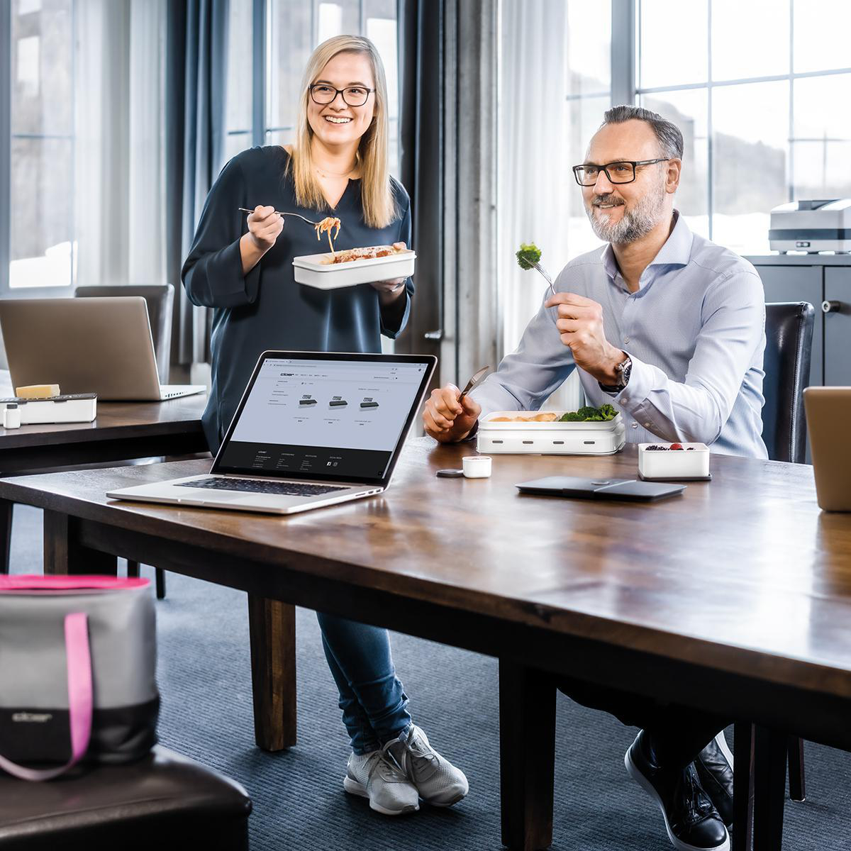 Zwei Personen essen Mittagessen an einem Holztisch, mit Laptops, Lebensmittelbehältern und einer grauen Tasche.