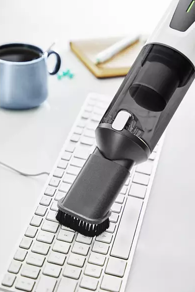 Ein Handstaubsauger reinigt eine Tastatur. Eine Kaffeetasse, ein Stift und ein Notizblock sind im Hintergrund.
