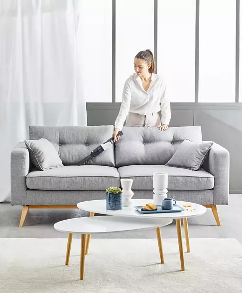 Frau saugt ein graues Sofa ab. Ein weißer Couchtisch mit Deko steht davor.