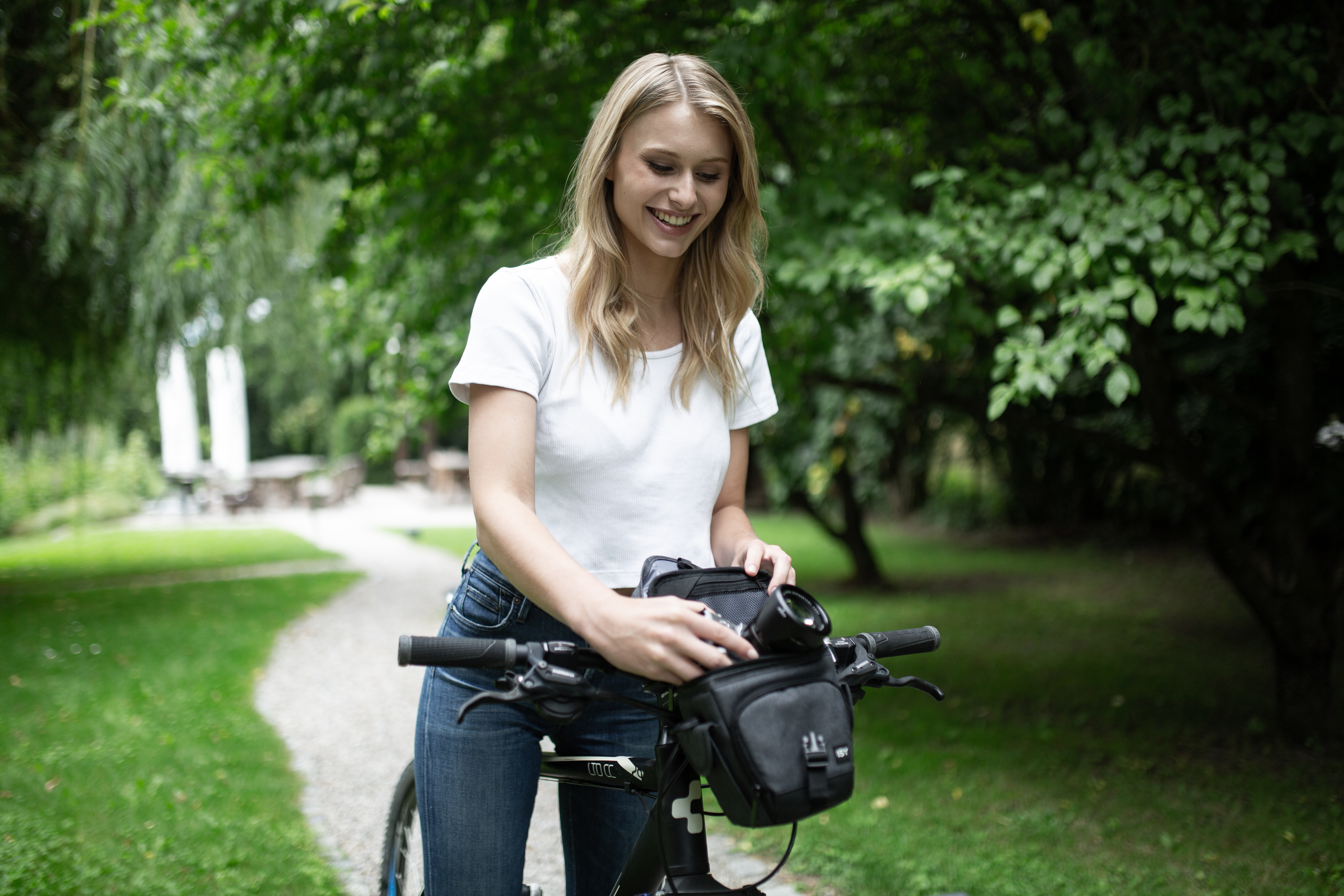 Eine Frau montiert eine Kameratasche an einem Fahrrad, lächelnd in einem Park.