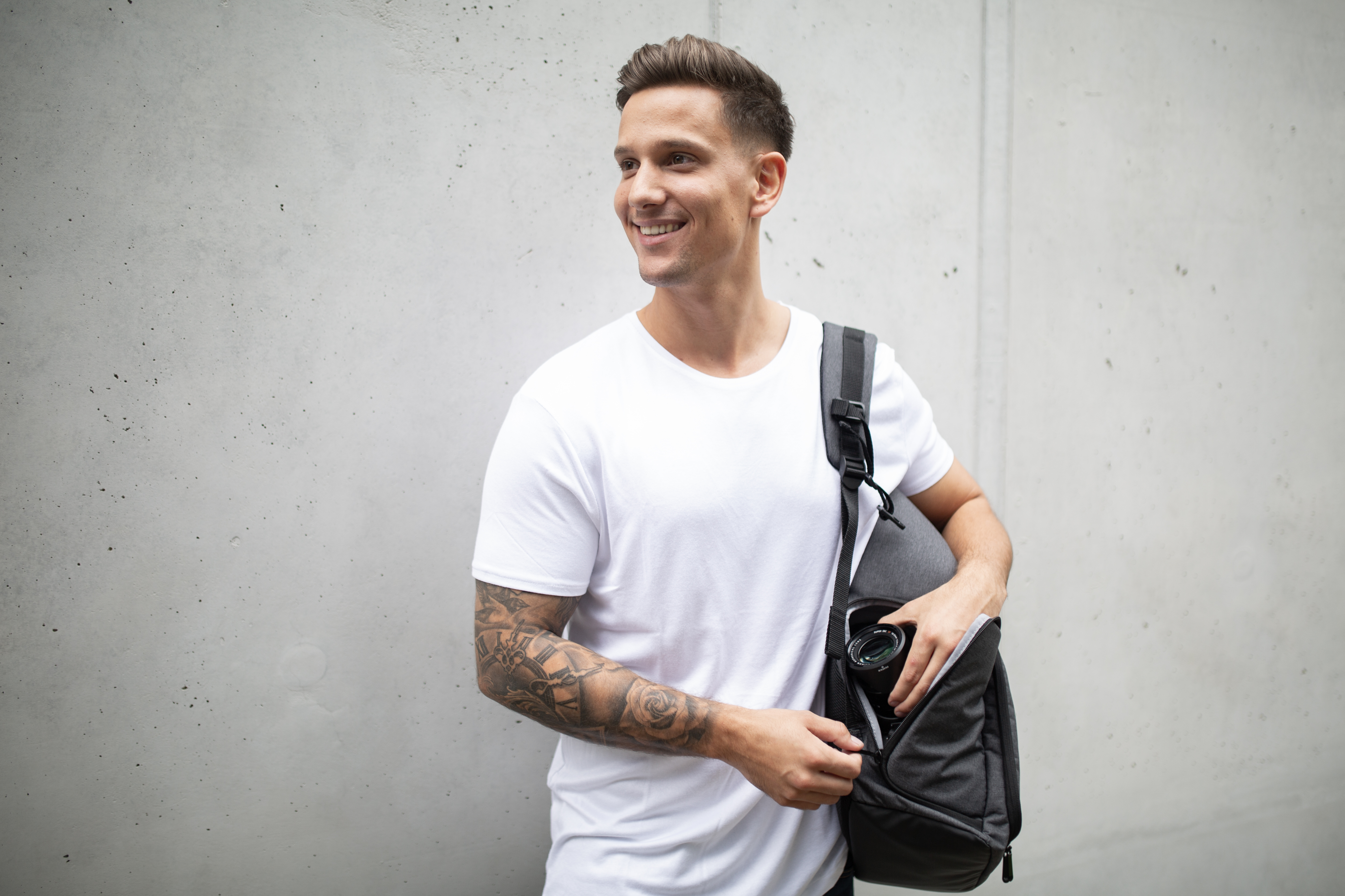 Ein Mann mit Tattoos und weißem Hemd lächelt und hält eine Tasche mit einer Kamera.