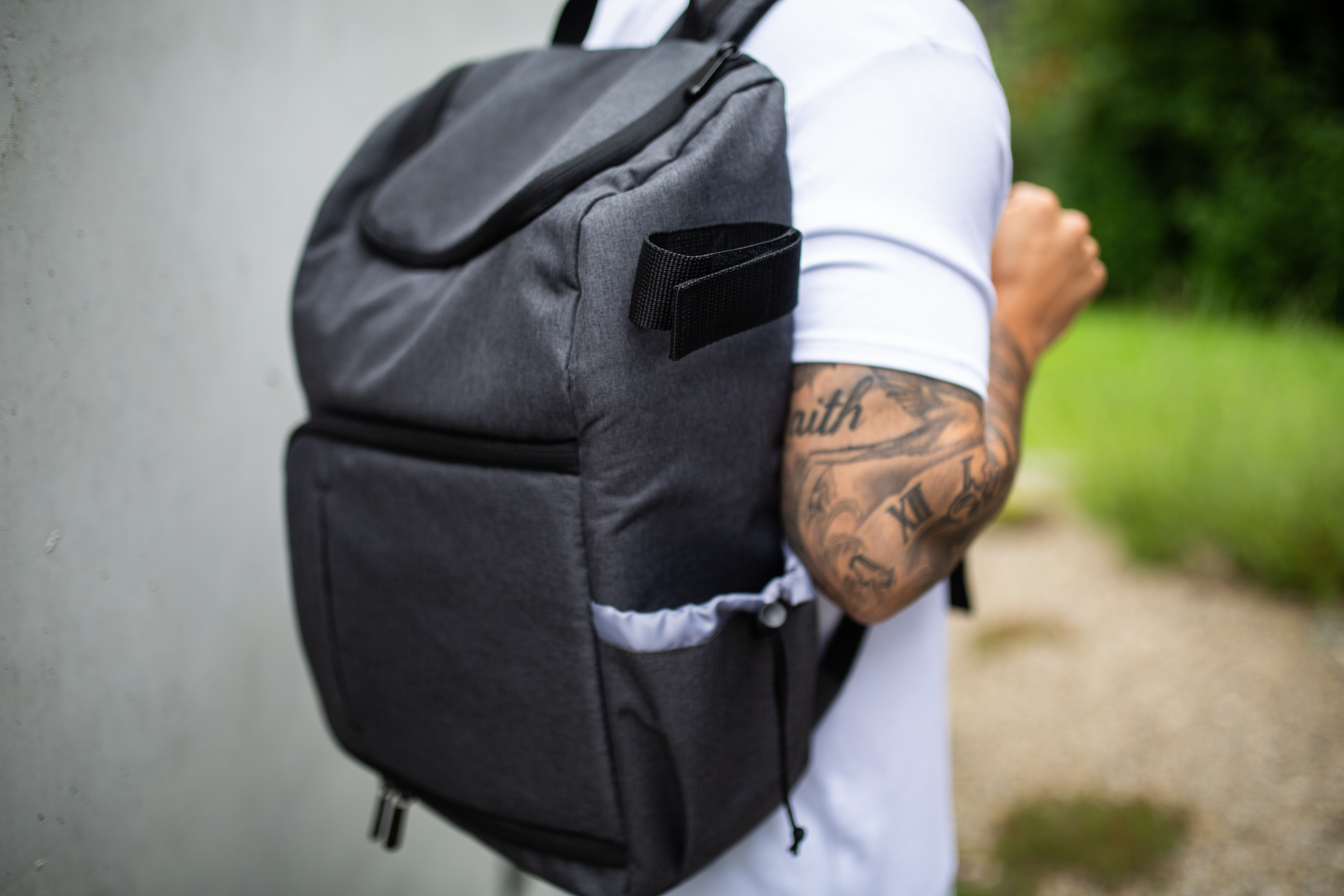 Eine Person mit Tätowierung am Arm trägt einen dunkelgrauen Rucksack mit schwarzem Gurt.