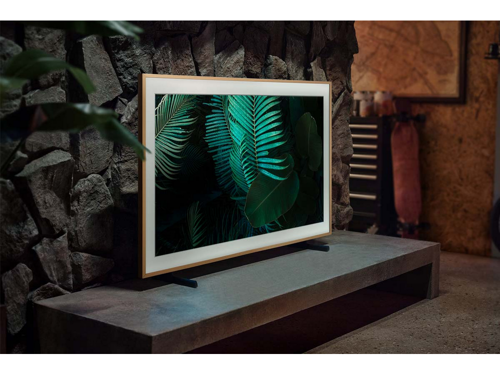 Un televisor moderno sobre un soporte, con un fondo de pared de piedra y hojas verdes.