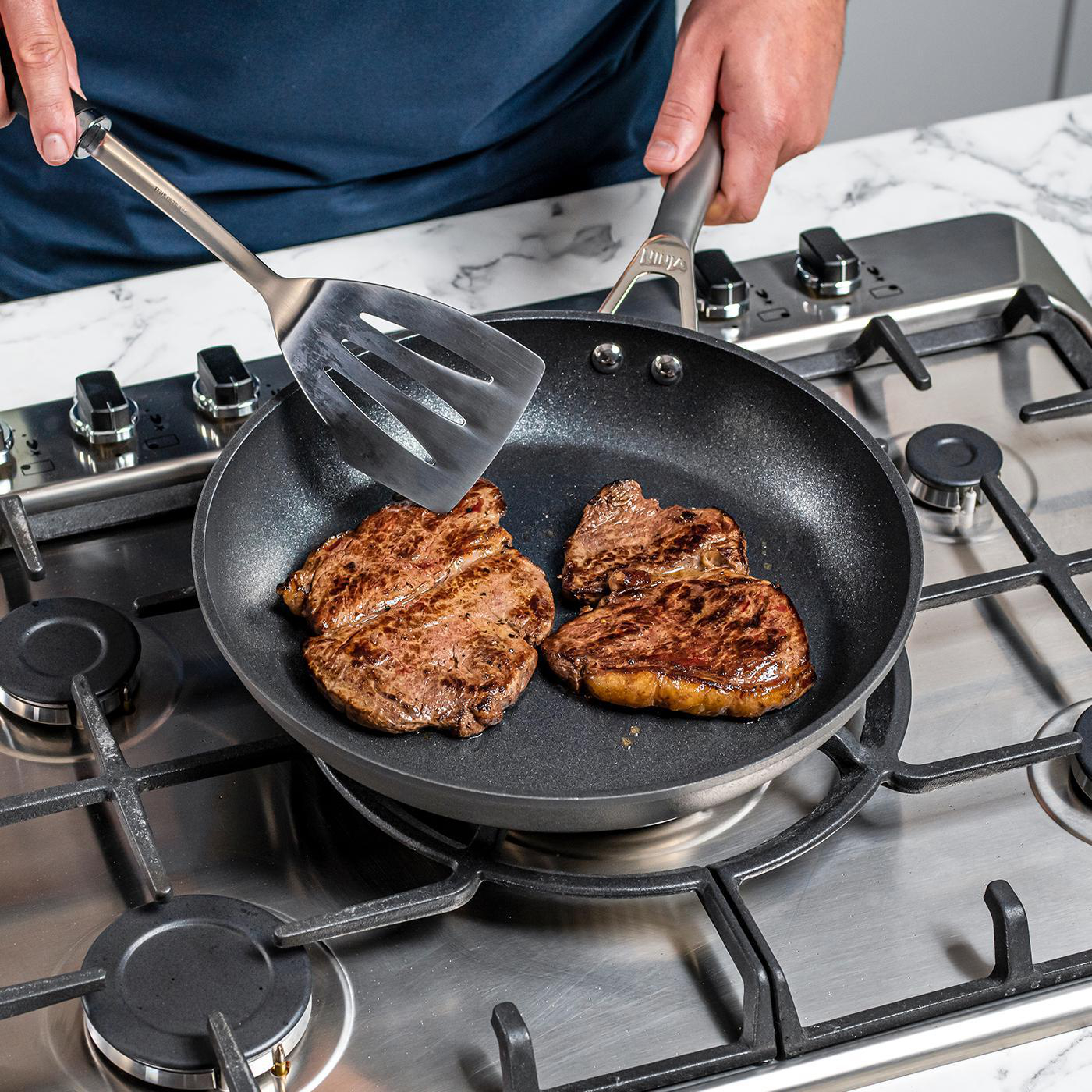 Person wendet Steaks in einer Bratpfanne auf einem Gasherd. Drei gebratene Steaks sichtbar.