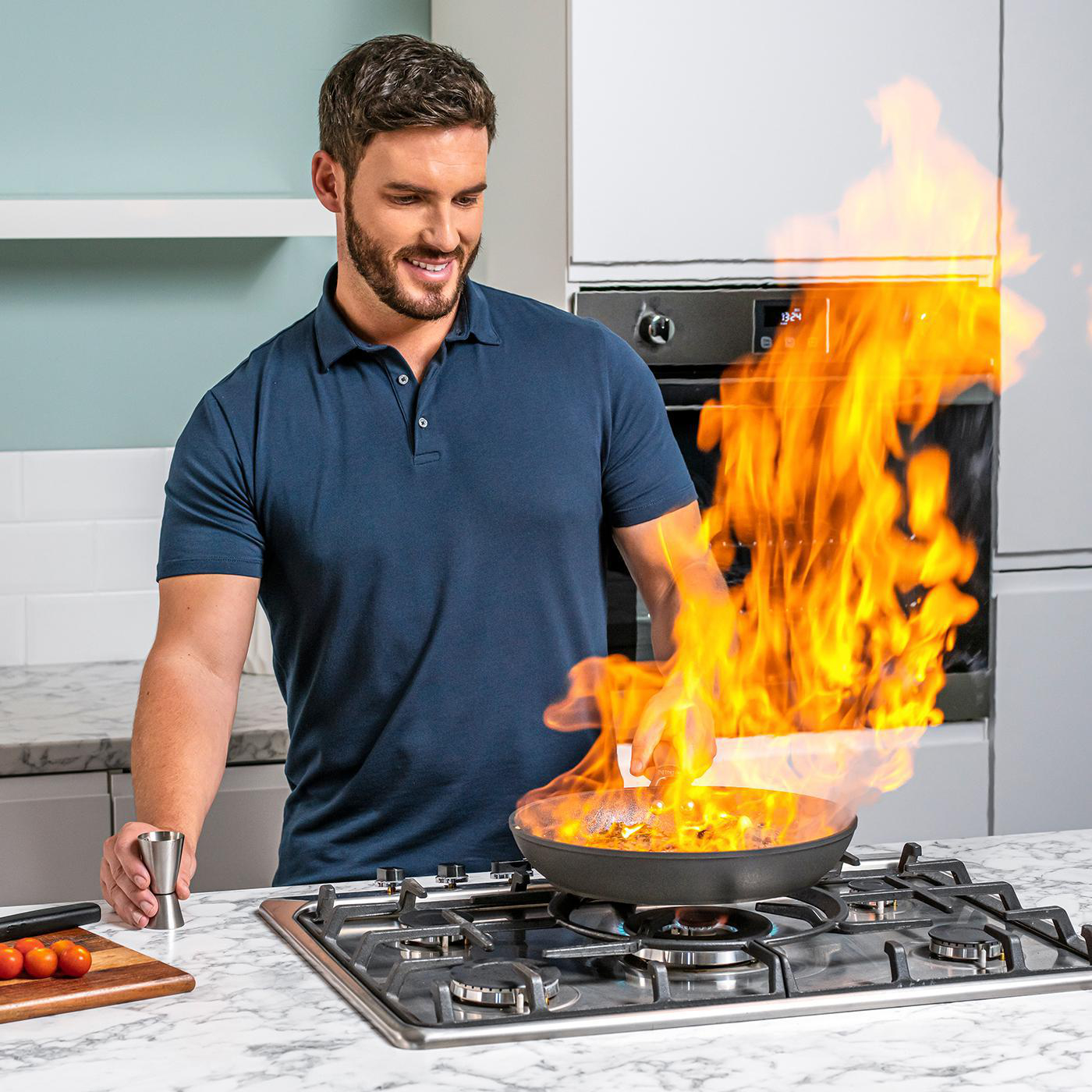 Ein Mann kocht mit Feuer in einer Pfanne auf einem Herd, mit einem Messbecher und Tomaten.