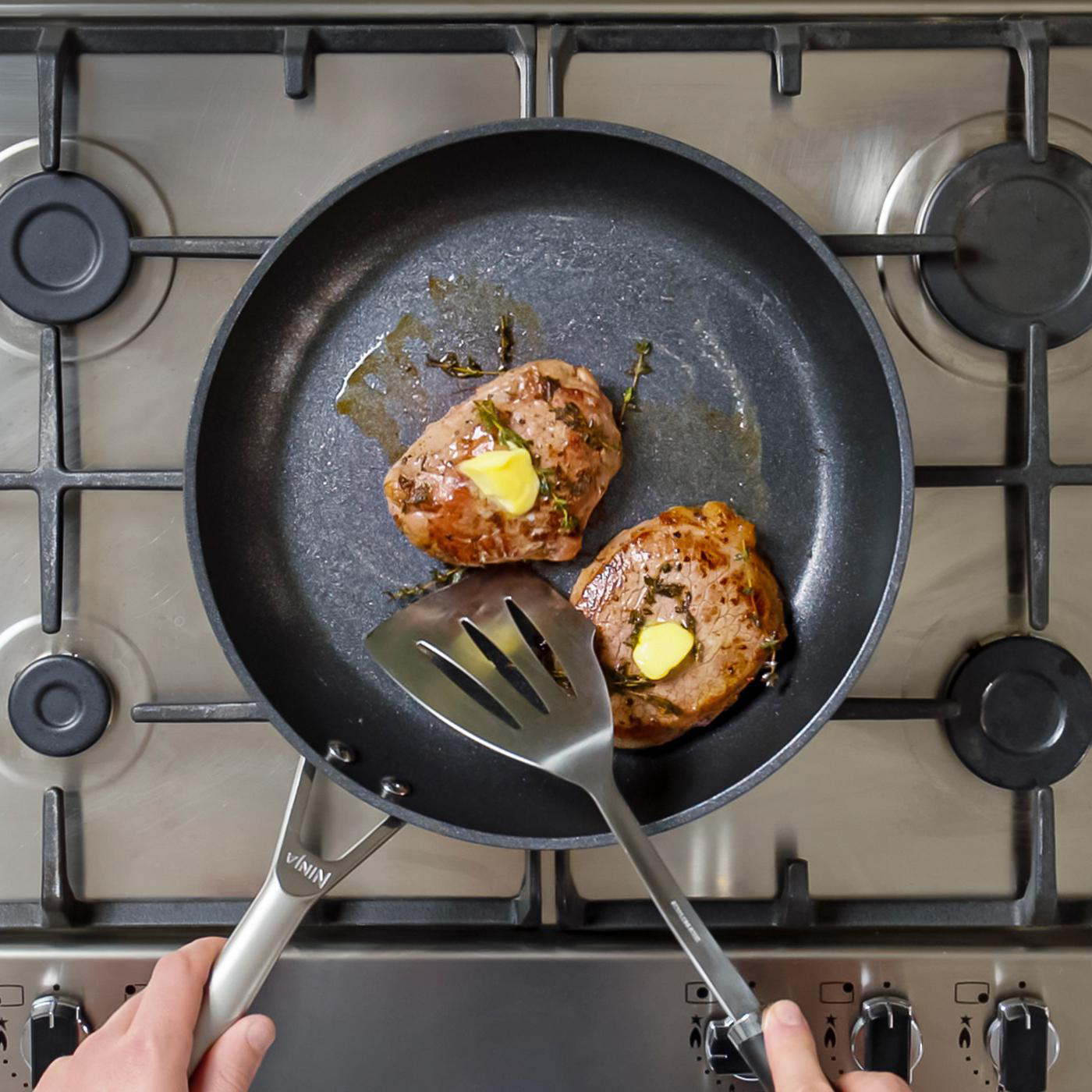 Zwei Steaks in einer Pfanne mit Butter und Kräutern, gehalten von einem Pfannenwender und einem Griff.