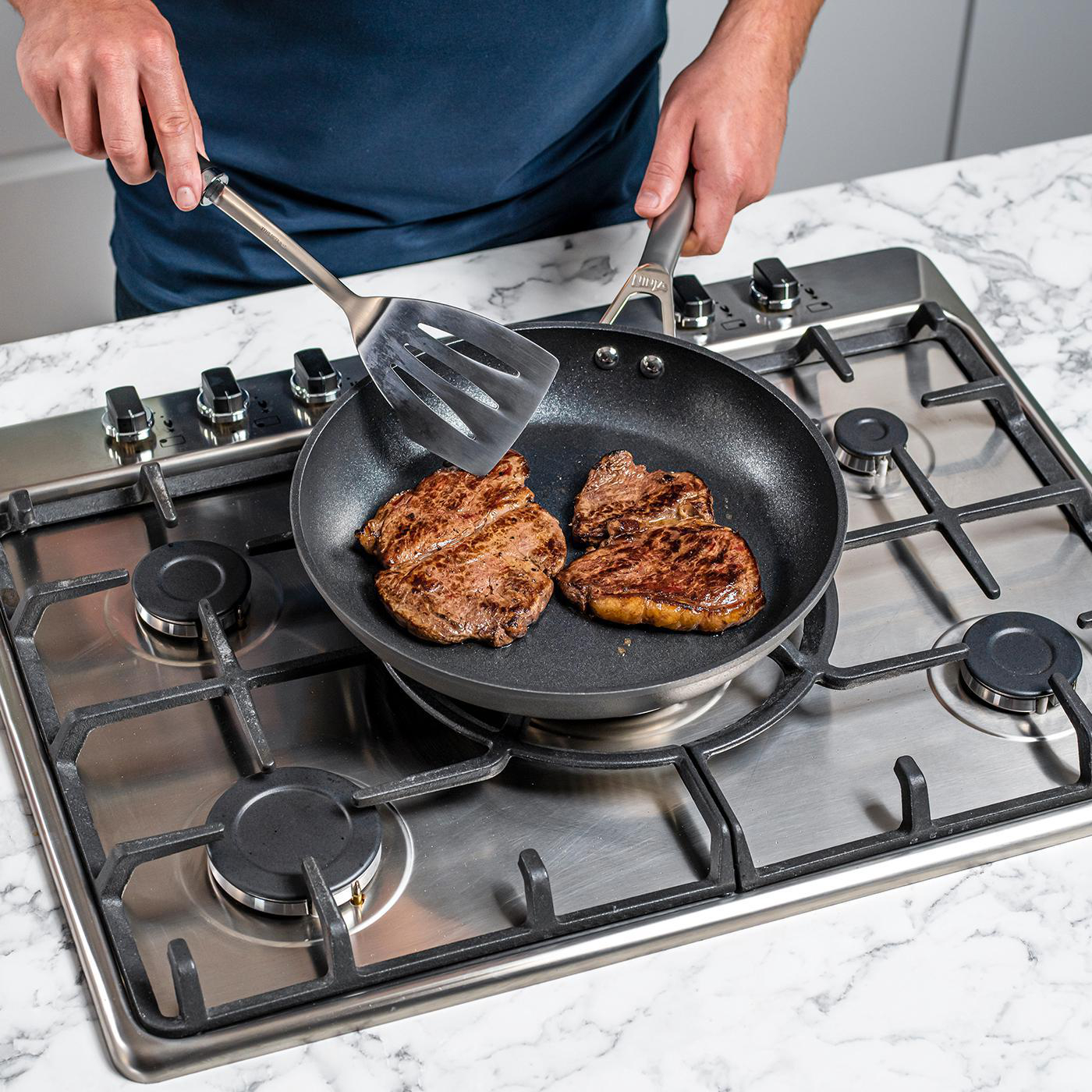 Ein Mann in blauem Hemd brät Steaks in einer Pfanne auf einem Gasherd.