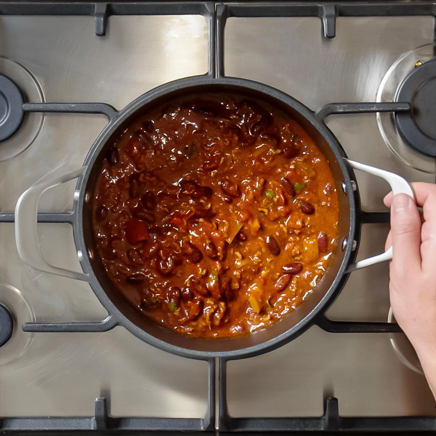 Eine Pfanne mit Chili con Carne steht auf einem Herd. Eine Hand hält die Pfanne.