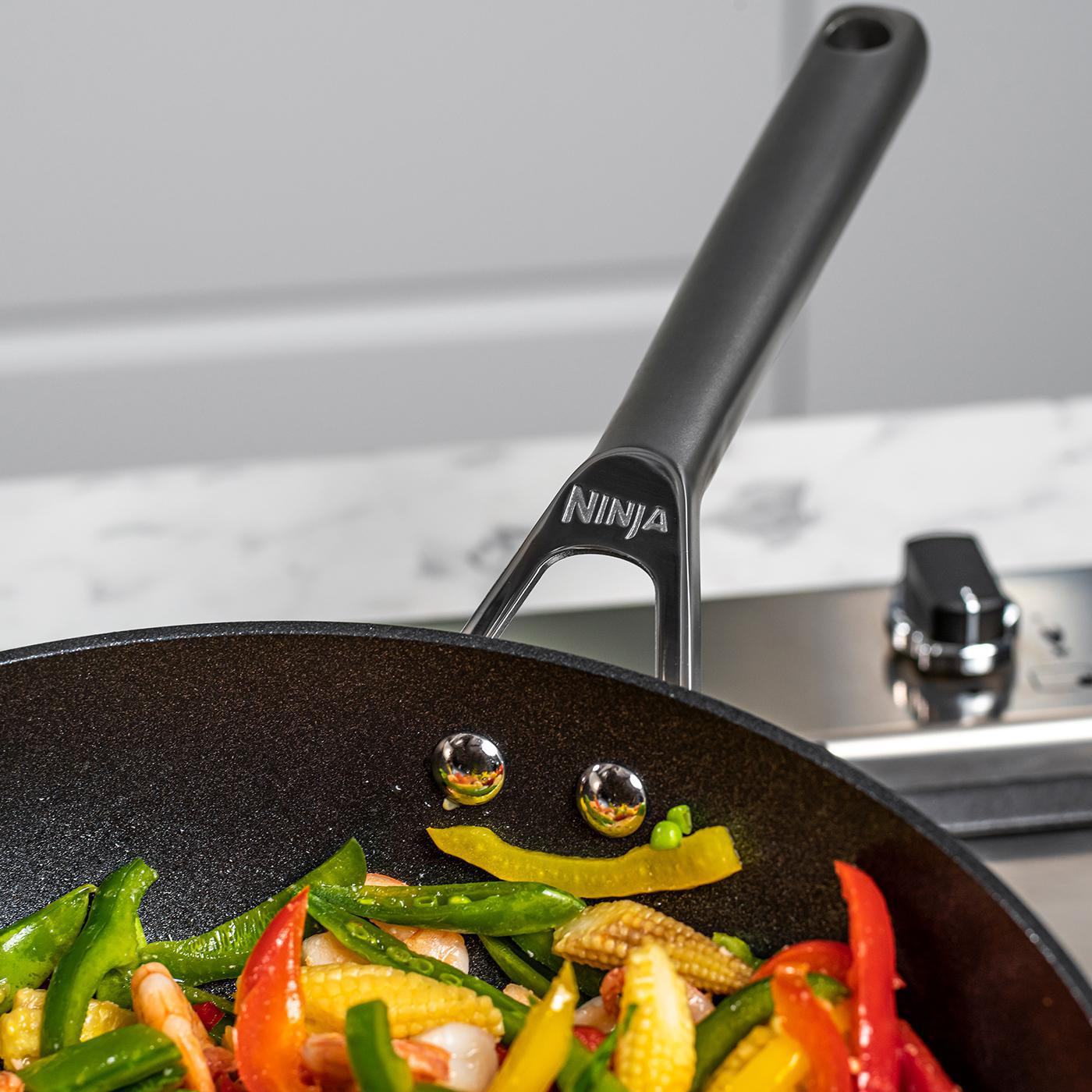 Nahaufnahme einer Bratpfanne mit Gemüse und Garnelen beim Kochen. Schwarze Pfanne, Ninja-Logo.
