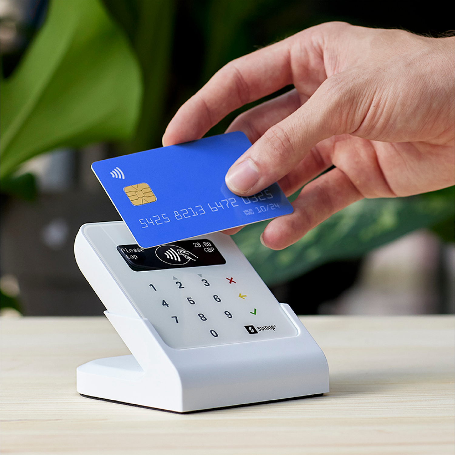 A hand holding a blue credit card over a white payment terminal. The terminal displays a screen.