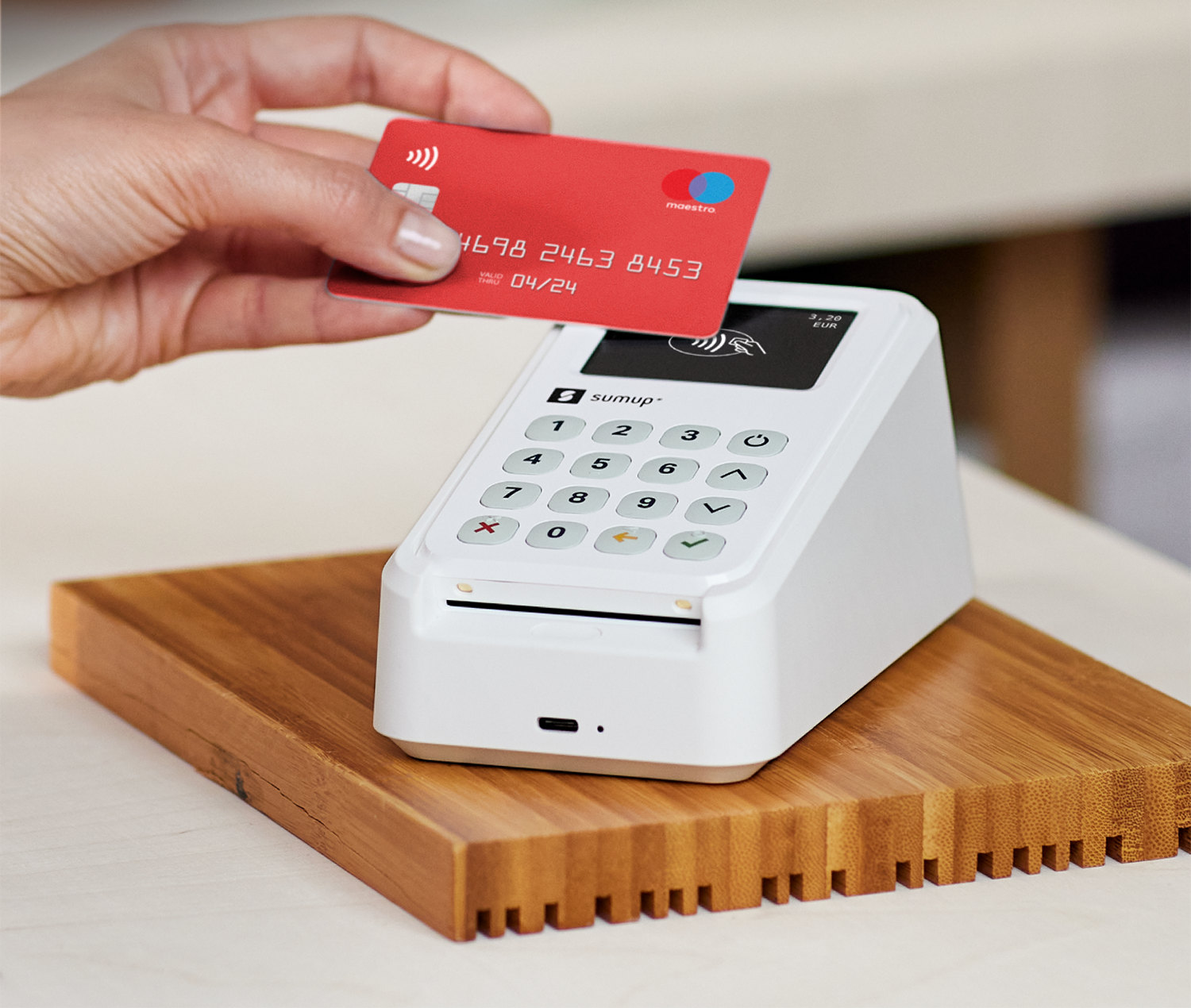 Hand holding a red credit card over a white payment terminal on a wooden base.