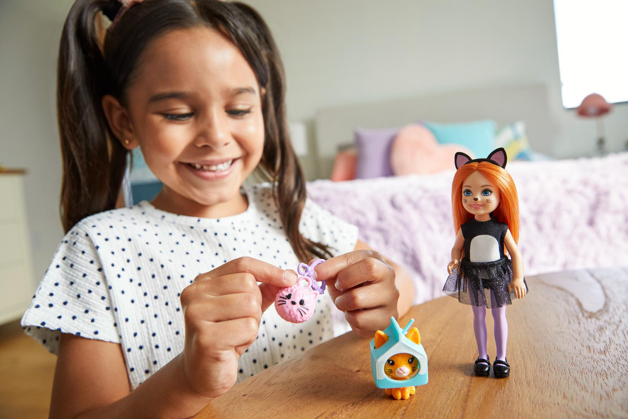 Ein Mädchen lächelt eine Katzpuppe und Spielzeug an. Die Puppe trägt Katzenohren und ein Kleid.