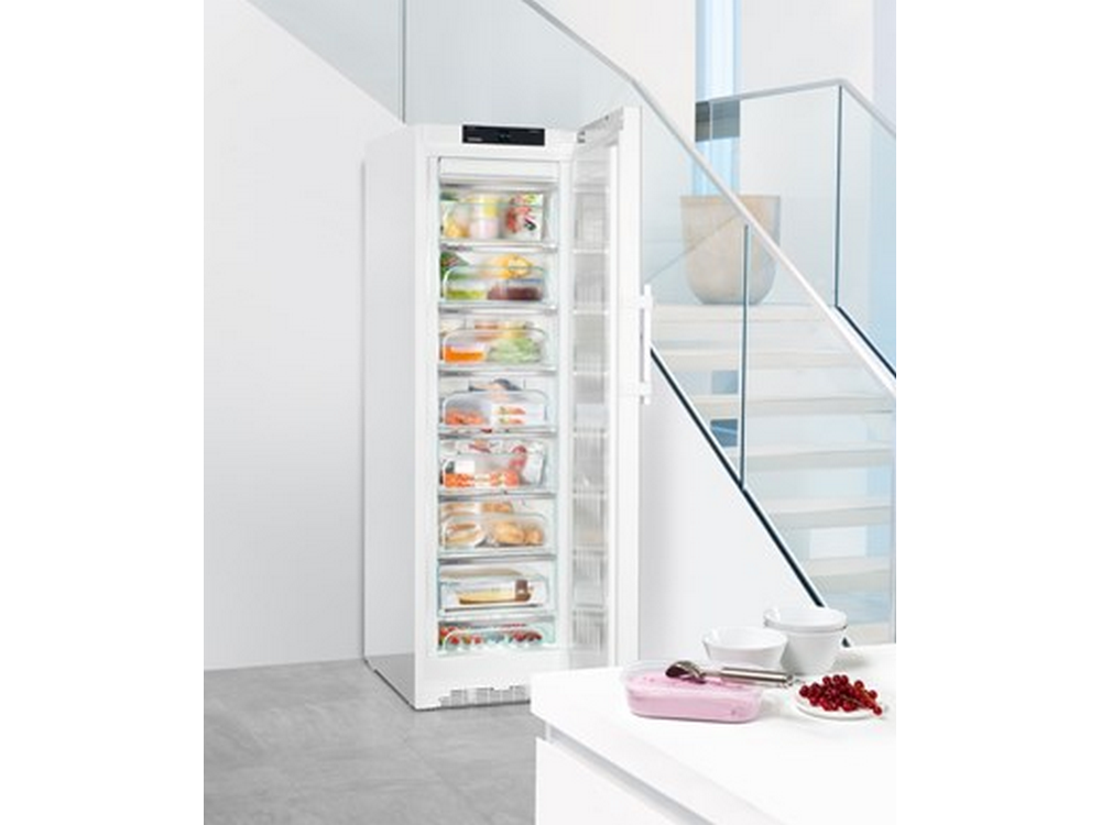 Refrigerador blanco con la puerta abierta. Dentro hay estantes con comida. Escaleras y pared blanca al fondo.