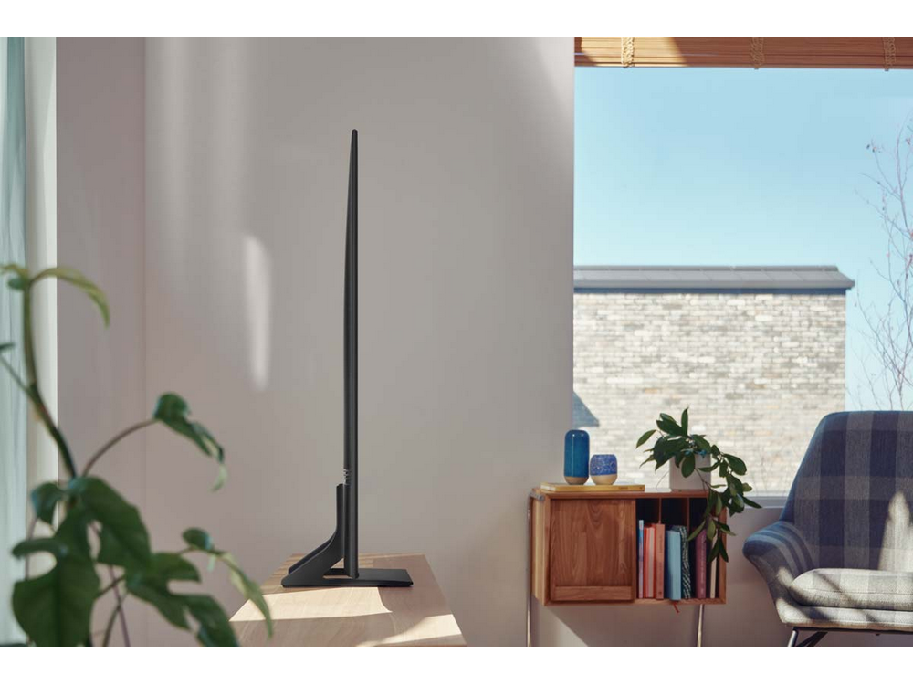 Un televisor negro moderno y delgado está sobre una mesa de madera. Una ventana y plantas están en el fondo. La habitación es luminosa.