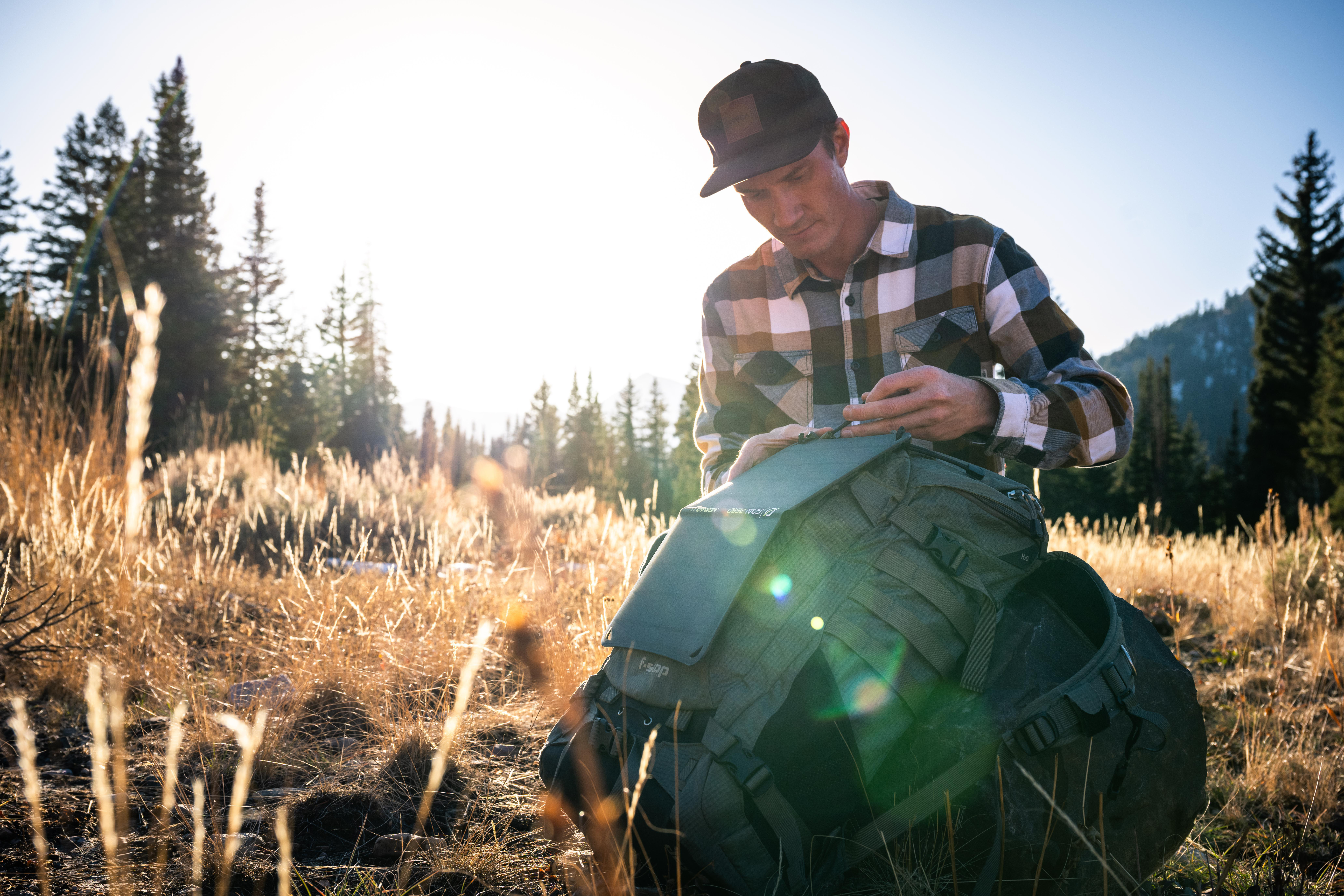 Ein Mann im karierten Hemd öffnet einen Rucksack auf einem Feld. Die Sonne scheint durch die Bäume im Hintergrund.