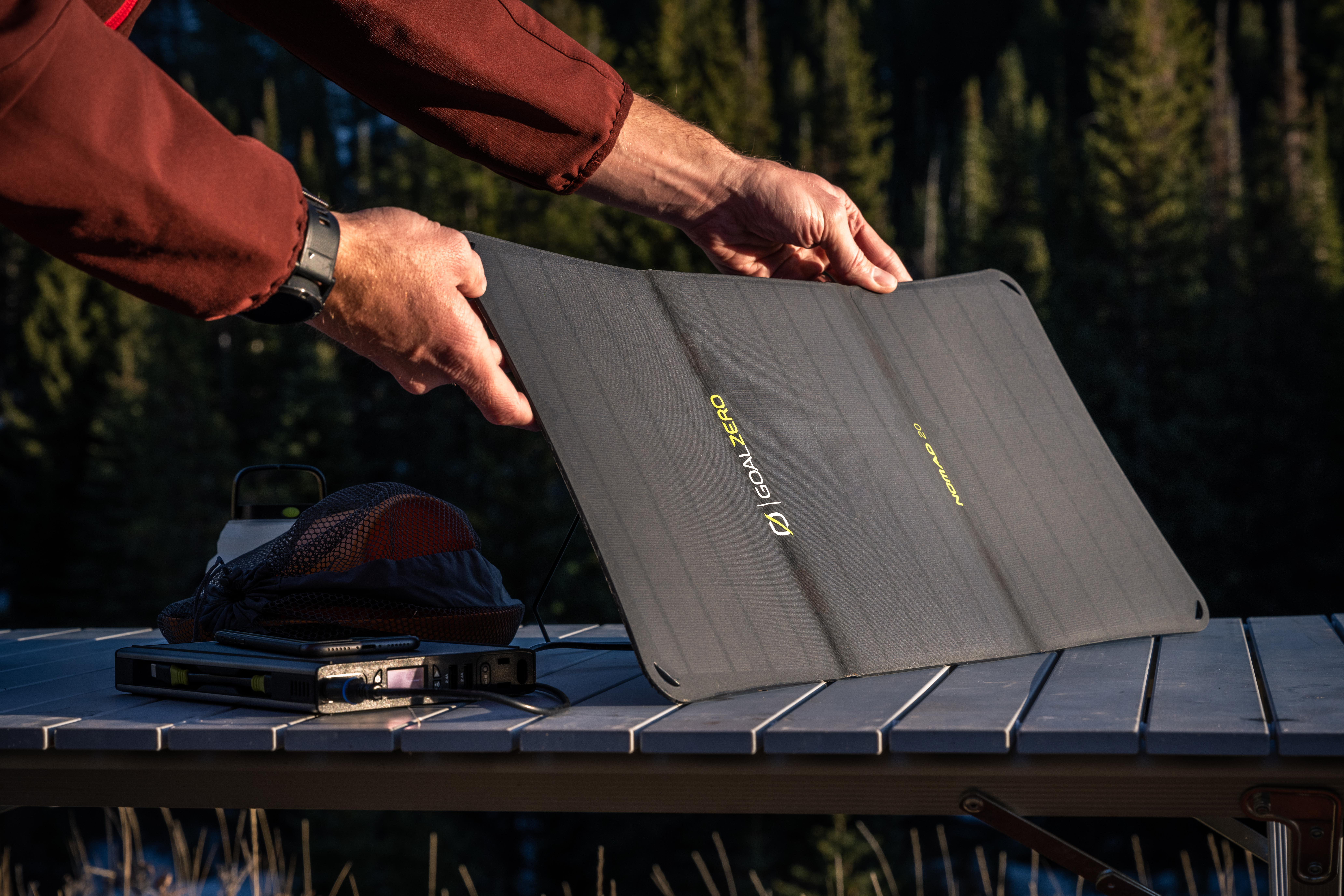Eine Person hält ein schwarzes Solarpanel auf einem Tisch. Andere Gegenstände wie eine Batterie und ein Ladegerät sind sichtbar.