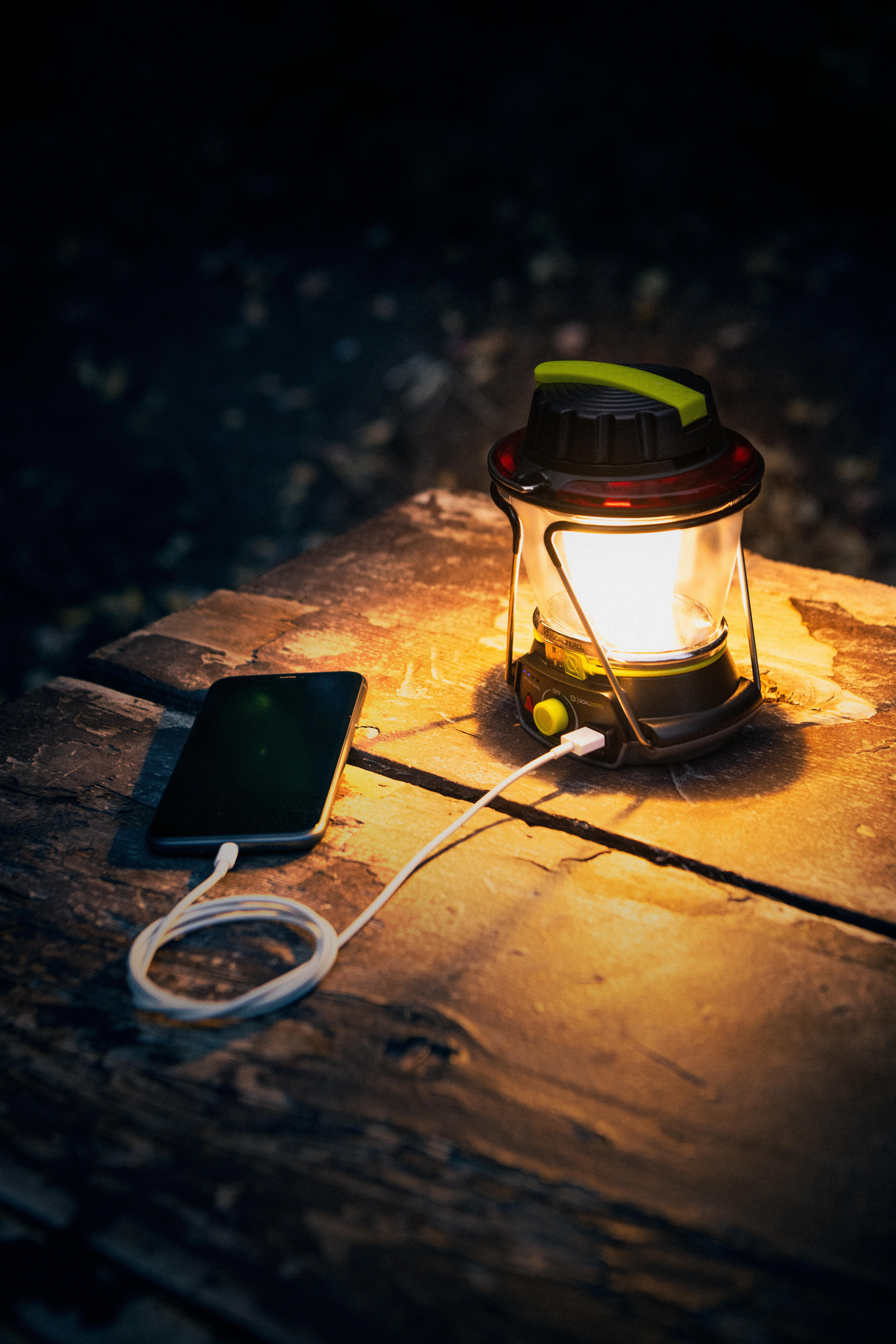 Eine beleuchtete Laterne und ein Handy, das mit einem weißen Kabel auf einem Holztisch bei Nacht geladen wird.