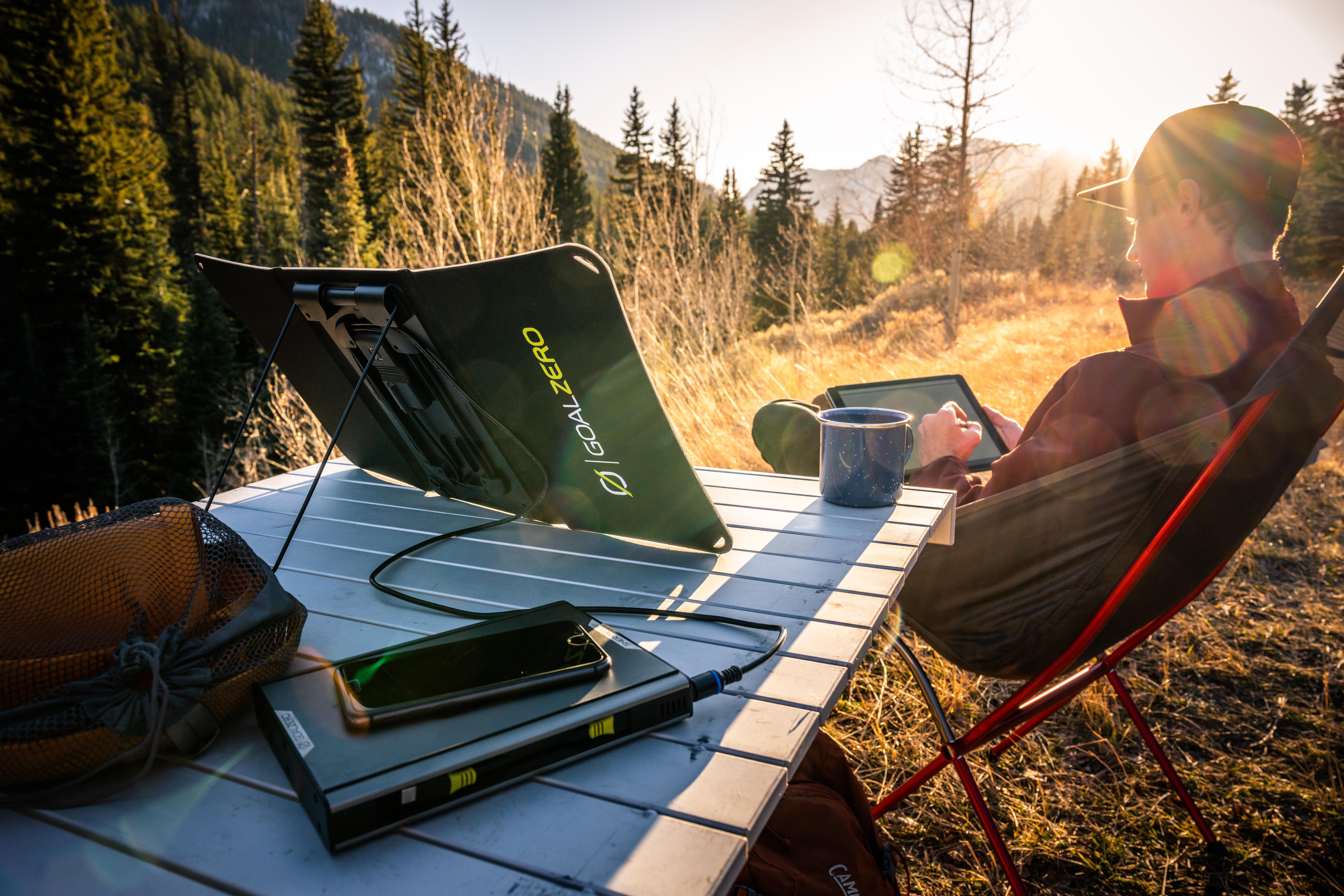 Eine Person benutzt ein Tablet an einem Tisch mit Solarpanel und Campingausrüstung in der Natur.