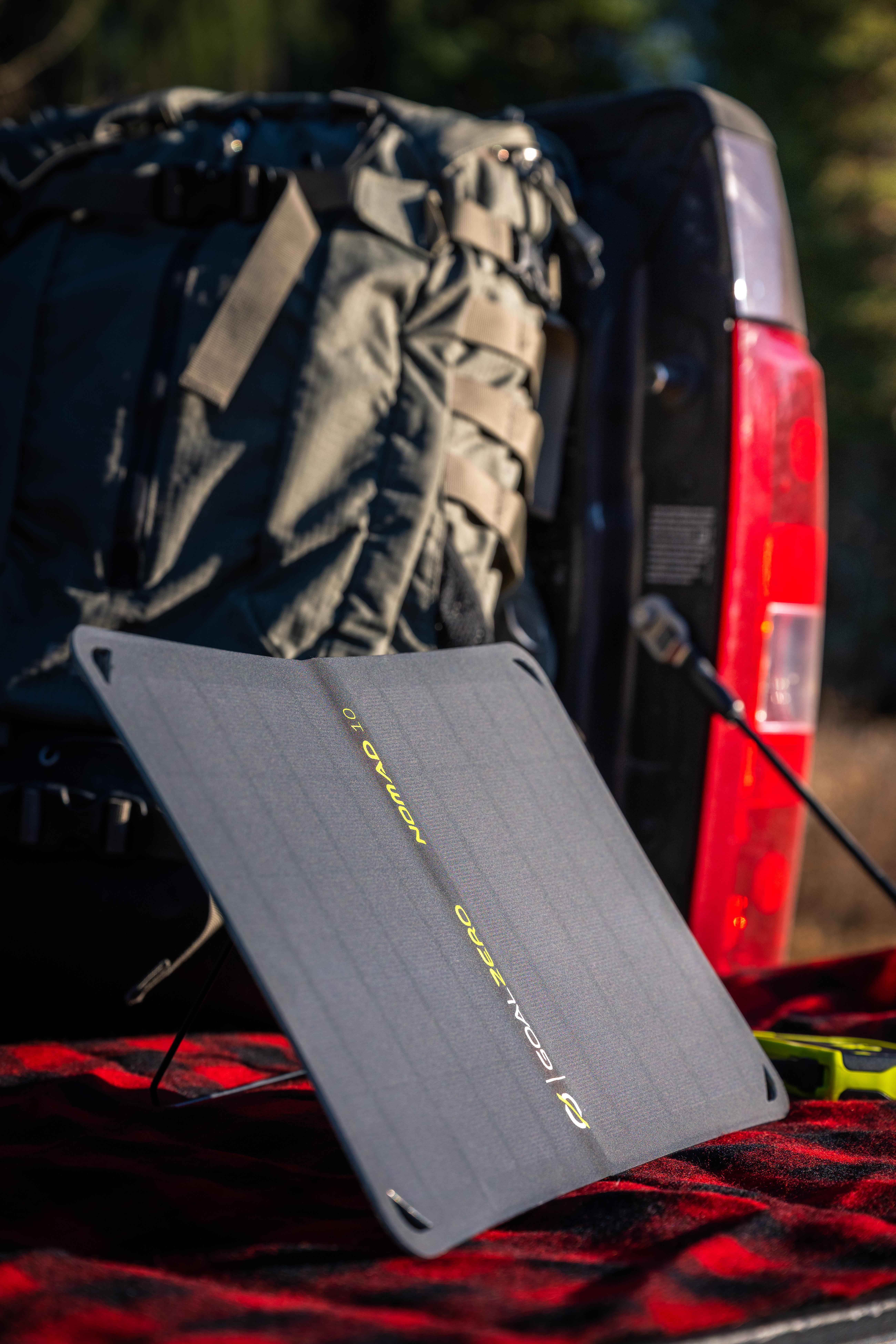 Solarpanel auf rot-karierter Oberfläche mit Rucksack, LKW und Bäumen im Hintergrund.