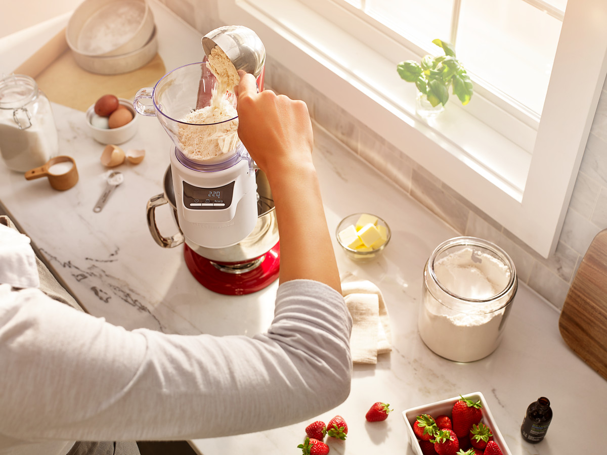 Eine Person gießt Mehl in eine rote Küchenmaschine, daneben Erdbeeren und Butter auf der Theke.