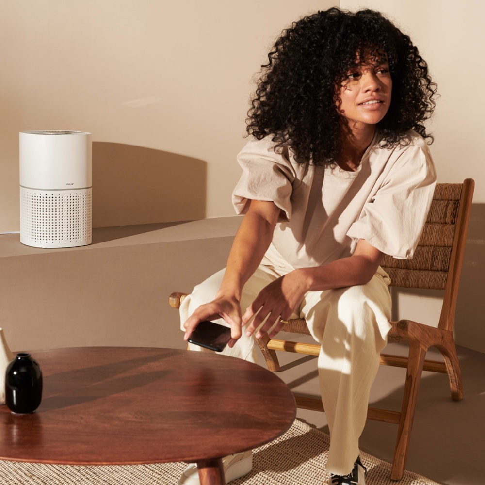 Een vrouw met krullend haar zit op een houten stoel en houdt een telefoon vast. Er staat een ronde tafel met een vaas en een luchtreiniger op de achtergrond.