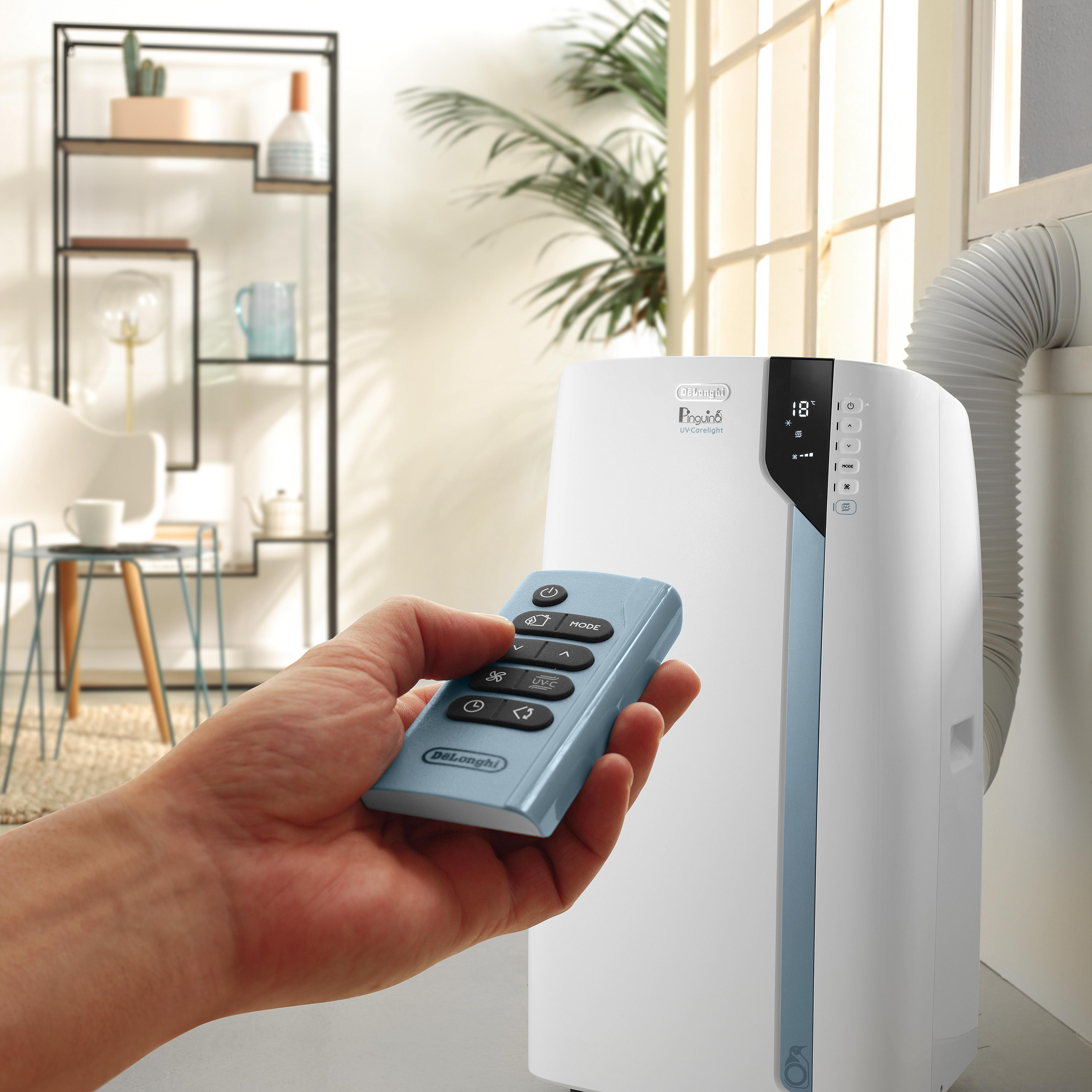 Hand, die eine hellblaue Fernbedienung hält, zeigt auf eine weiße mobile Klimaanlage.