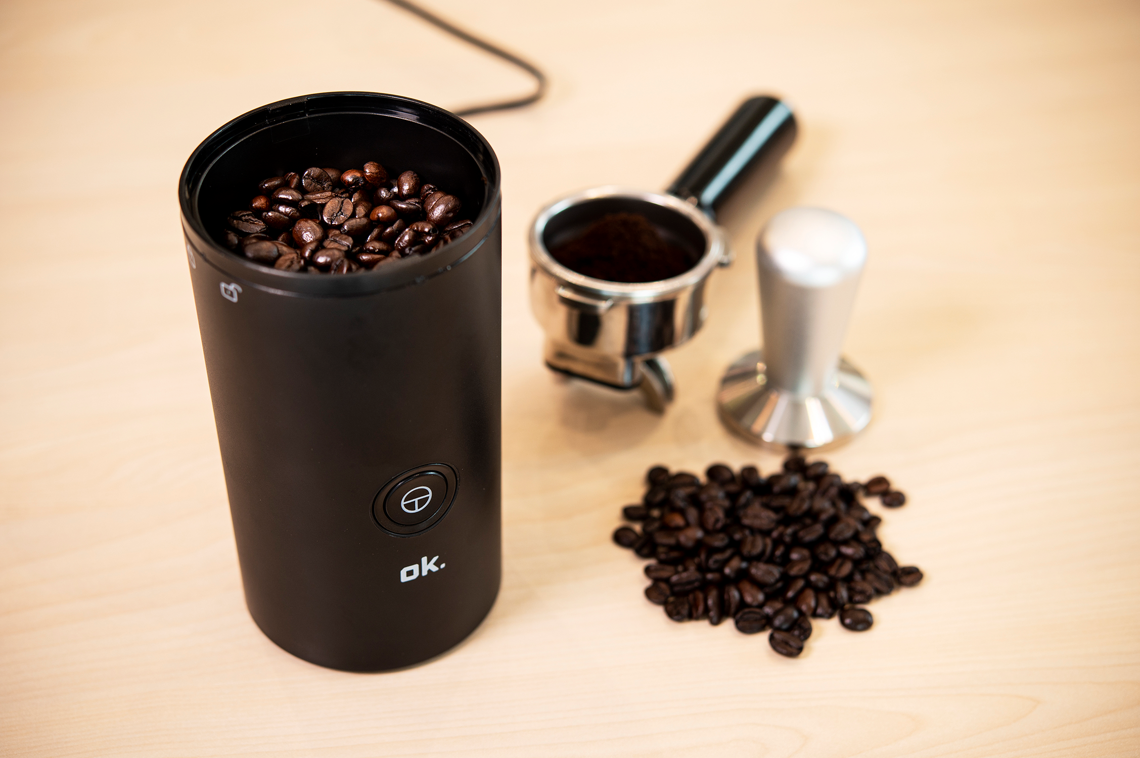 Coffee grinder with beans, a portafilter with coffee, and a tamper on a wooden table.
