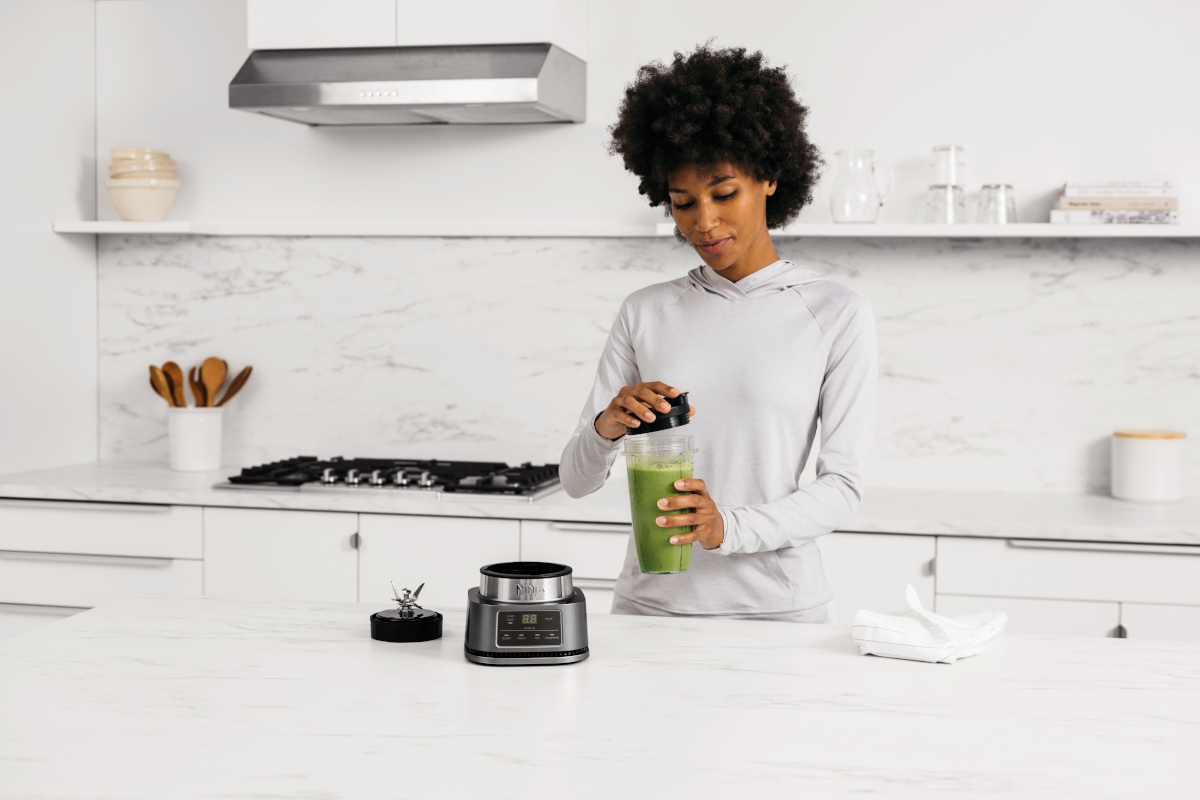 Woman in grey hoodie blending a green smoothie. Blender and ingredients are on a counter.
