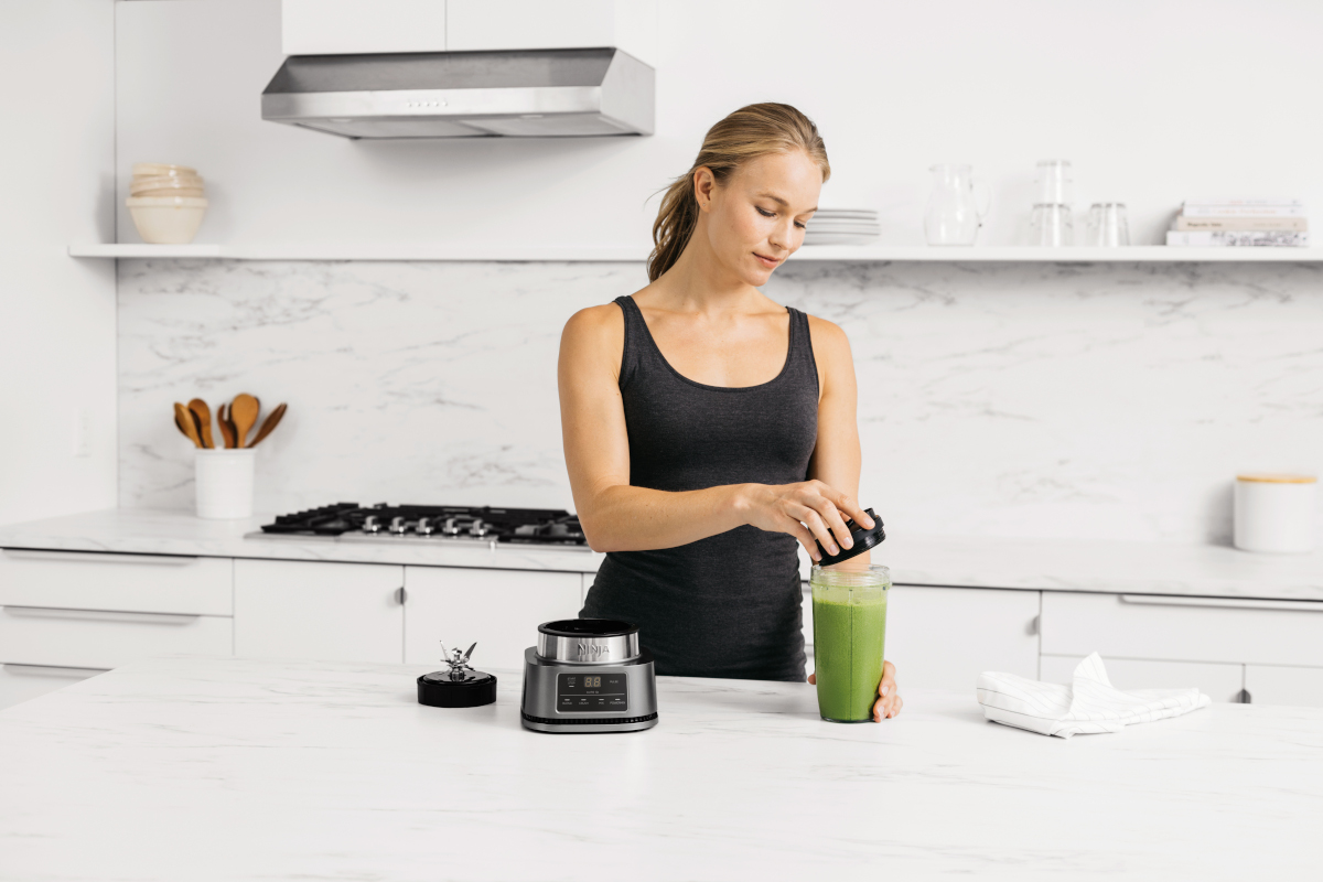 Woman in kitchen using a blender, with ingredients visible.