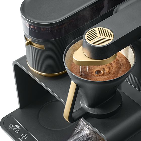 Machine à café, design noir et doré, en cours d'utilisation, le café coule dans une carafe foncée.