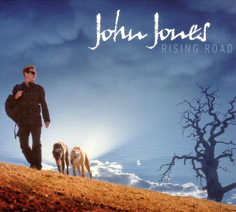 Ein Mann mit Rucksack und zwei Hunde gehen auf einem grasbewachsenen Hügel. Text: John Jones Rising Road.