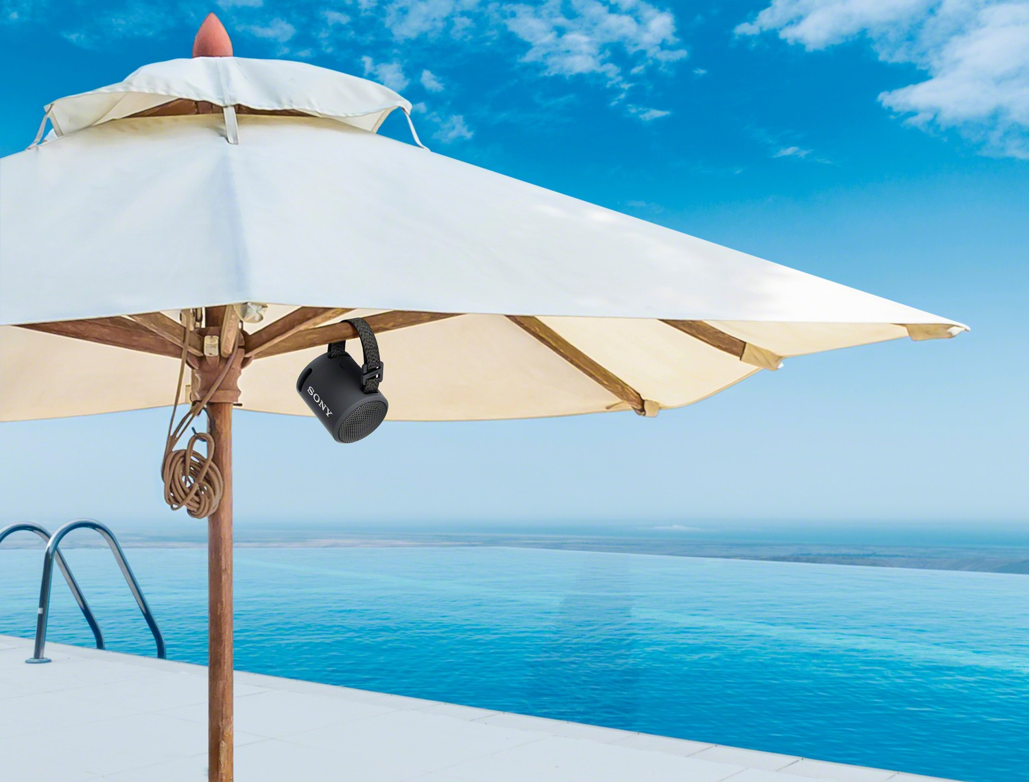 Un altavoz negro cuelga de una sombrilla blanca. Un piscina y el océano son visibles en el fondo.